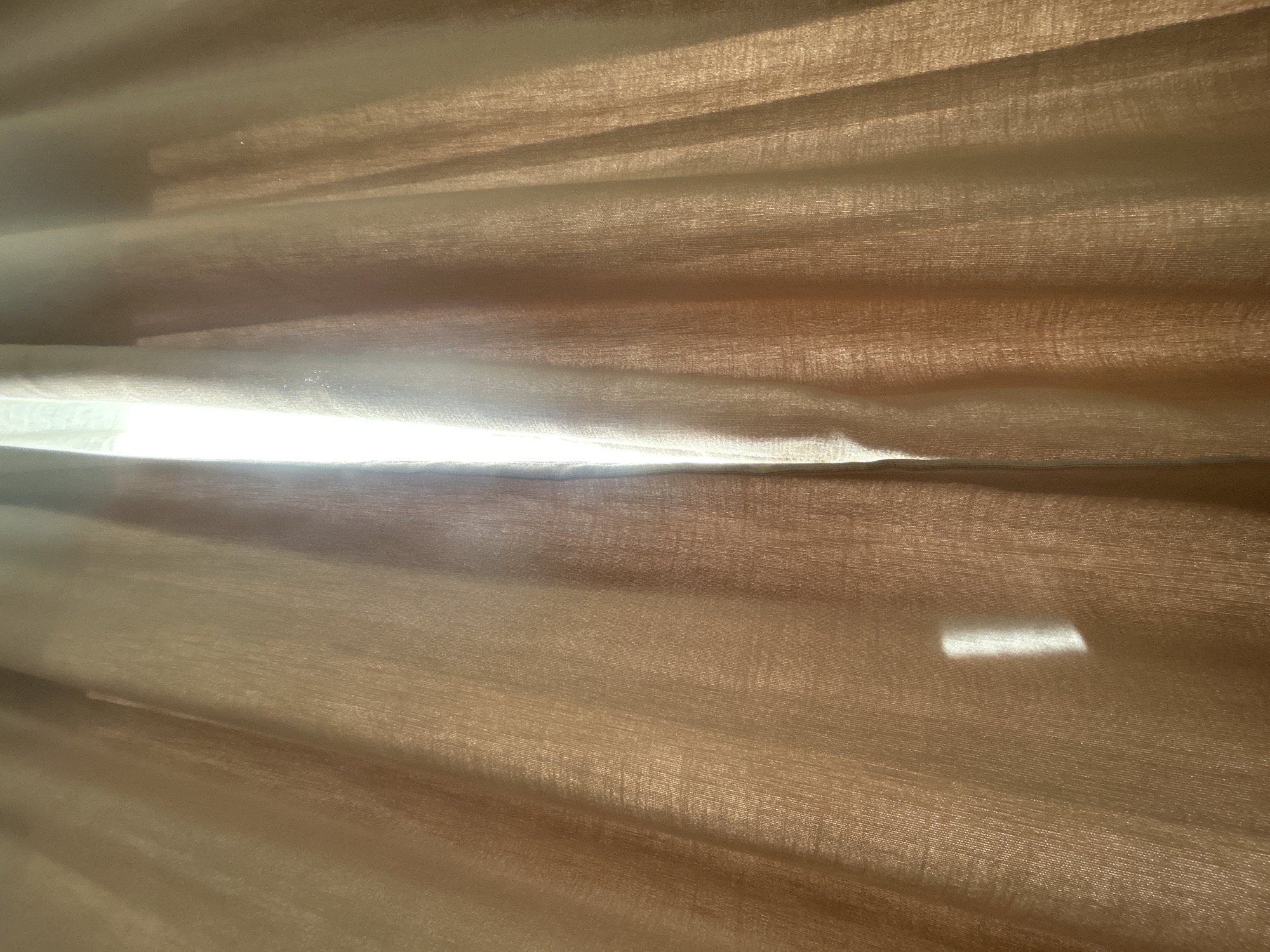 The image shows a close-up view of translucent, light brown curtains with vertical folds. Sunlight streams through a gap in the center of the curtains, creating a bright white vertical area. A rectangular reflection of light is visible on the left curtain. The scene depicts the interior of a room where light is filtering through window coverings. There are no people or specific landmarks visible to determine a location in Plovdiv, Bulgaria.