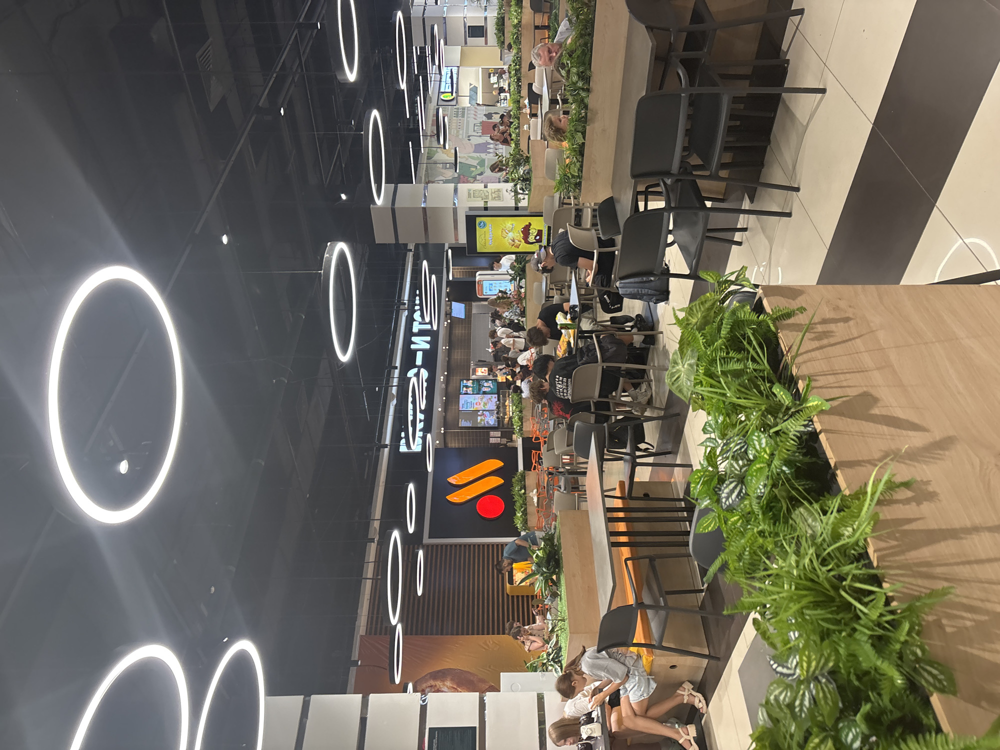 The image shows the interior of a food court, likely at a shopping mall. In the foreground, there are several tables and chairs, with a wooden planter box filled with lush green plants in front of them. In the midground, people are seated at various tables, eating and talking. There are large circular lights hanging from the ceiling, illuminating the space. Behind the food court area, there is a prominent sign with orange and red branding, suggesting a fast-food restaurant. The overall impression is a busy, modern dining environment. Based on the branding visible (the orange arcs), this is likely a McDonald's restaurant located in Naberezhnye Chelny, Russia, as the restaurant chain operated under the brand "Vkusno i tochka" (which translates to "Tasty and that's it") after it ceased operations in Russia.
