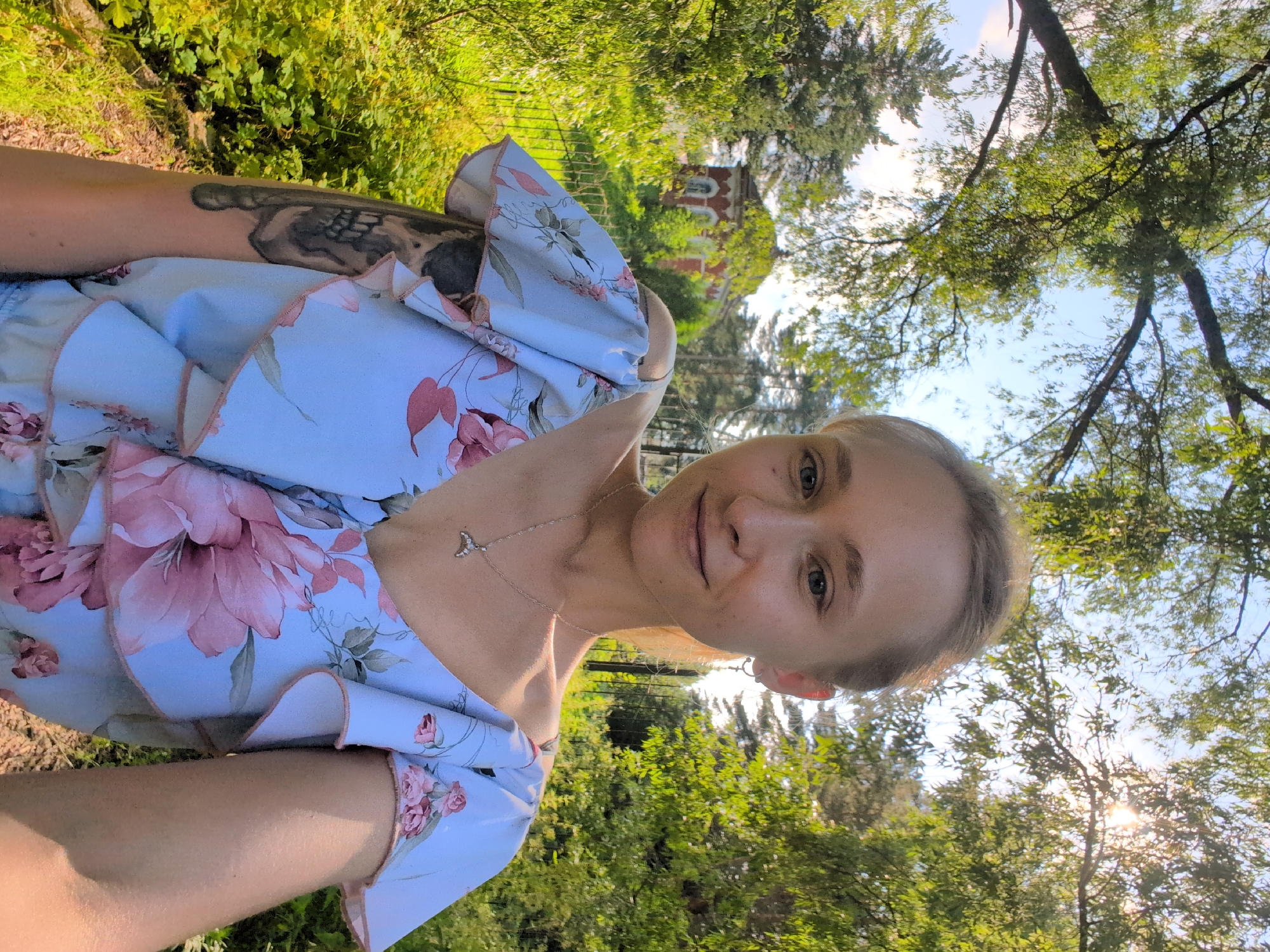 A woman with blonde hair and blue eyes is captured in a close-up, selfie-style photograph. She is wearing a light blue dress adorned with a floral pattern of pink flowers. A delicate silver necklace with a small pendant is visible around her neck. She has a tattoo on her left arm. The background suggests an outdoor setting with abundant green foliage, tall trees, and a clear blue sky, indicating a sunny day. Sunlight filters through the leaves, creating bright patches and lens flare. In the distance, a building with red brick and arched windows can be partially seen, hinting at a park or garden environment. The image is oriented sideways. The overall mood appears relaxed and pleasant, consistent with a warm, clear day spent outdoors. The context provided indicates the location as Veliky Novgorod, Russia.