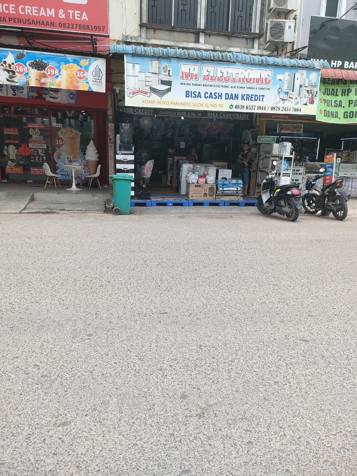 The image shows a street scene in Batam, Indonesia. On the left, a storefront with red signage advertises "ICE CREAM & TEA" with a phone number. Next to it, a storefront for "MR. ELECTRONIC" displays various electronic appliances and furniture. The signage also indicates "BISA CASH DAN KREDIT" (Can buy with Cash and Credit) and provides contact information. In front of the stores, there are several motorcycles parked, and a green trash bin is visible on the sidewalk. A person is standing in the doorway of the electronics store. The street is paved, and there are buildings with windows and air conditioning units in the background.