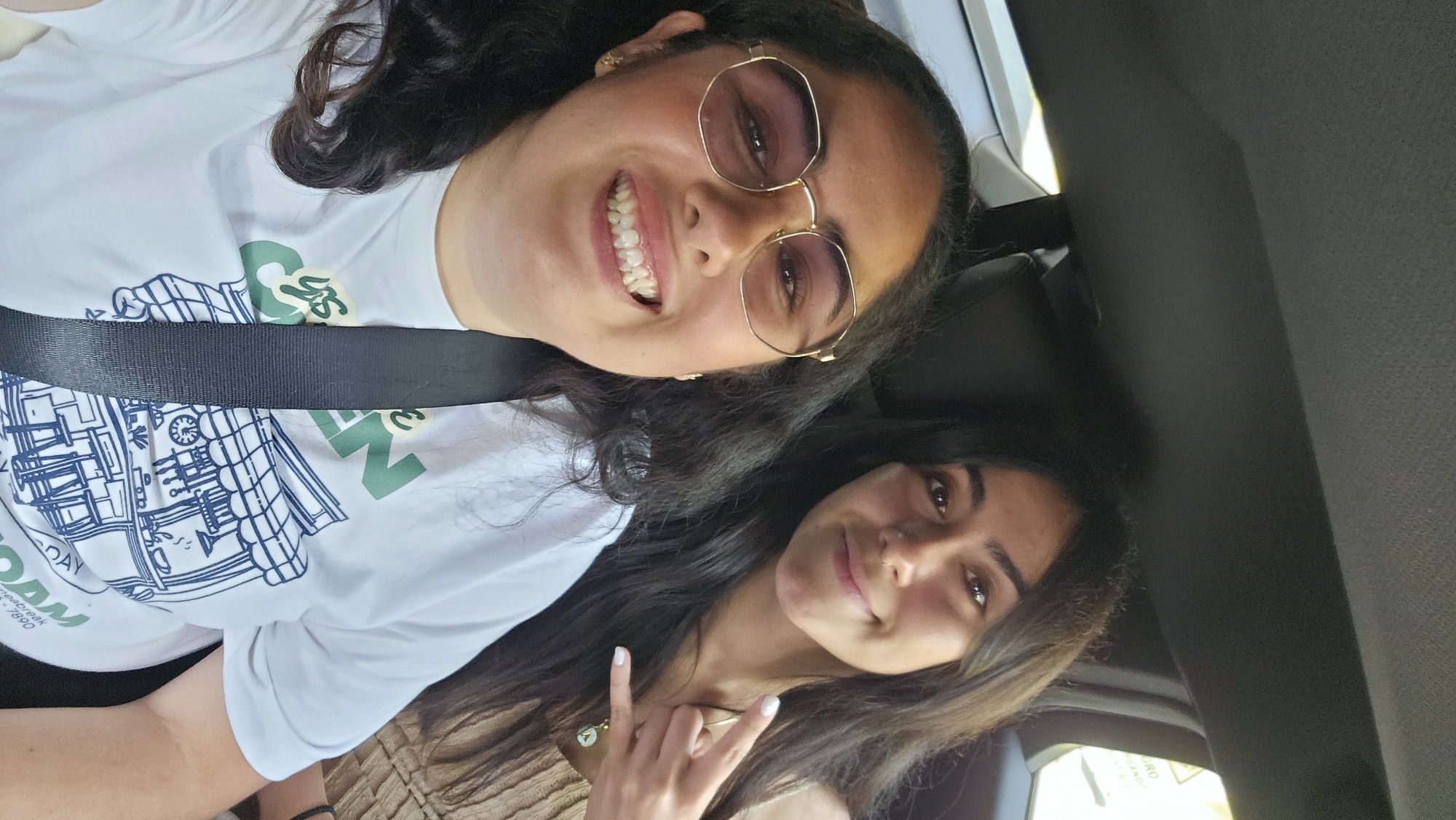 Two women are seated in what appears to be a car, possibly in Mexico City, Mexico. The woman in the foreground is wearing eyeglasses and a white t-shirt with a graphic design. A black seatbelt is visible across her torso. She is smiling broadly. The second woman, positioned in the background, is also smiling and making a "rock on" hand gesture. She is wearing a necklace. The interior of the car is visible, with black upholstery and parts of the car's structure. The lighting is bright, suggesting it is daytime.