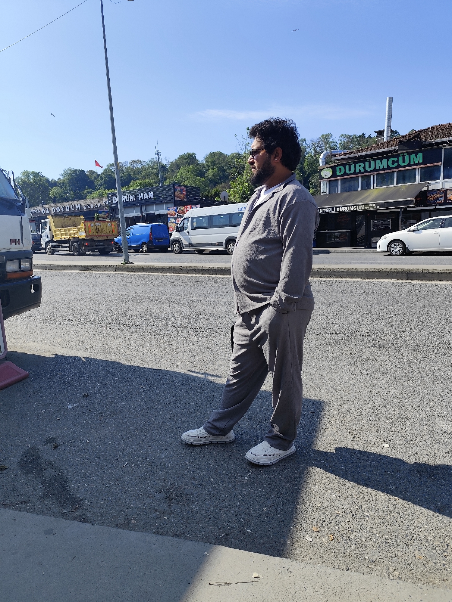 A man with dark hair and a beard stands on a street in Sarıyer, Turkey. He is wearing a gray suit and white shoes, with his right hand in his pocket. Behind him, there is a street with a variety of vehicles, including a truck, a bus, and a car. The "DÜRÜMCÜM" restaurant sign is visible in the background, along with a sign for "Döner Öz Doydum Kebab" and "Dürüm Han". The scene is illuminated by sunlight, casting a shadow from the man onto the street.