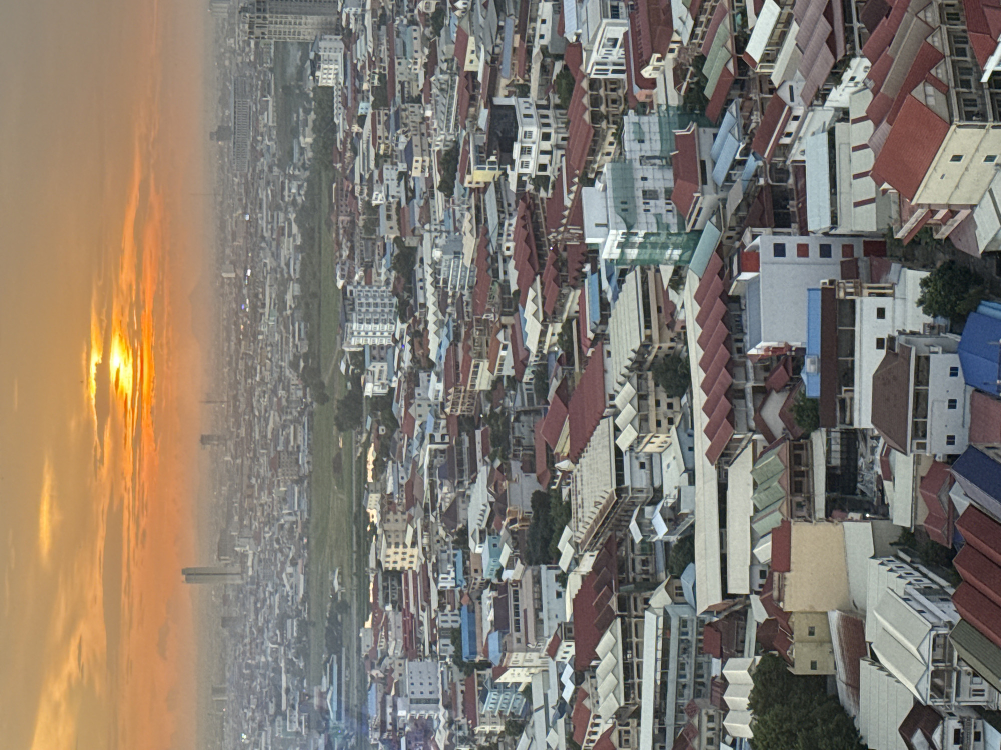 This is a high-angle, vertical, full-shot photograph capturing an expansive view of a dense urban cityscape at sunset, with the sky dominated by a warm orange and yellow glow. The sun is partially visible through hazy clouds, casting a soft light over the scene. The city appears to be located in Khan Mean Chey, Cambodia, indicated by the architectural styles and density of buildings.
The landscape is characterized by a multitude of buildings, predominantly residential and commercial structures, packed closely together. Many buildings feature distinctively angled, multi-pitched red-tiled roofs, a common architectural feature in the region. The structures vary in height, with a mix of low-rise apartment buildings, multi-story houses, and a few taller, modern high-rise buildings scattered throughout the urban sprawl. Some buildings exhibit balconies, while others have flat roofs. Several structures are in various stages of construction or renovation, with some partially covered by green netting.
The urban environment is characterized by a lack of green space within the densely packed residential areas, with only small patches of trees visible between buildings. The overall impression is one of a rapidly developing city with a mix of traditional and contemporary architectural styles. No people or vehicles are clearly discernible due to the distance and the nature of the aerial perspective. The atmospheric conditions suggest a slight haze, common during sunset in urban areas. The photograph captures the vastness and density of the urban fabric under the dramatic lighting of the setting sun.