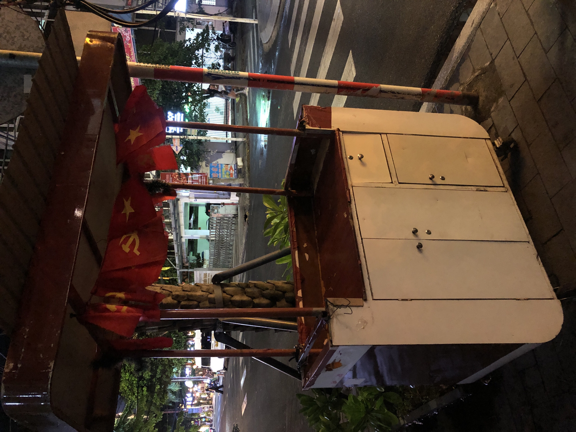 This media file shows a street scene at night in Da Nang, Vietnam. The primary focus is a food cart or stall with a white cabinet on the right side, featuring two drawers with silver knobs. A red metal frame supports a canopy or awning on the left, which is adorned with two Vietnamese flags, each featuring a yellow star on a red background. These flags appear to be made of fabric and are slightly tattered.
Behind the cart, a red and white striped barrier, likely a gate or bollard, is visible, extending horizontally across the scene. The ground is paved with rectangular stones on the right, and a wet asphalt road with a pedestrian crossing is visible on the left. Streetlights illuminate the scene, reflecting on the wet surfaces, suggesting recent rain or a humid environment. Lush green foliage is present in the background, adding a touch of nature to the urban setting.
Further back, buildings with illuminated signs and windows can be seen, indicating commercial activity. A few people are visible in the distance, blurred by the low light and motion, suggesting the street is active at night. The overall impression is a typical street view in a Southeast Asian city after dark, with elements of commerce, daily life, and potentially national pride represented by the flags. The time of day is clearly night, and the wet surfaces imply damp weather conditions. There is no discernible text that can be read clearly from the image.