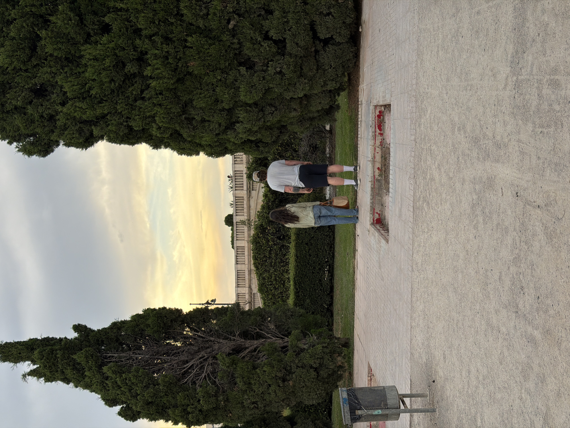 The image shows two people standing in a park in the city of València, Spain. The man, wearing a light-colored t-shirt and dark shorts, stands to the right. The woman, in a light-colored shirt and jeans, stands to his left. They are both facing away from the camera, looking at a landscaped area in the distance. The park features well-maintained green spaces and paved walkways. Two large, dark green evergreen trees frame the central view. In the background, a low, cream-colored building with arched windows is visible behind a neatly trimmed hedge. The sky above is a pale orange and yellow, suggesting either sunrise or sunset. A trash can is visible on the left side of the image.