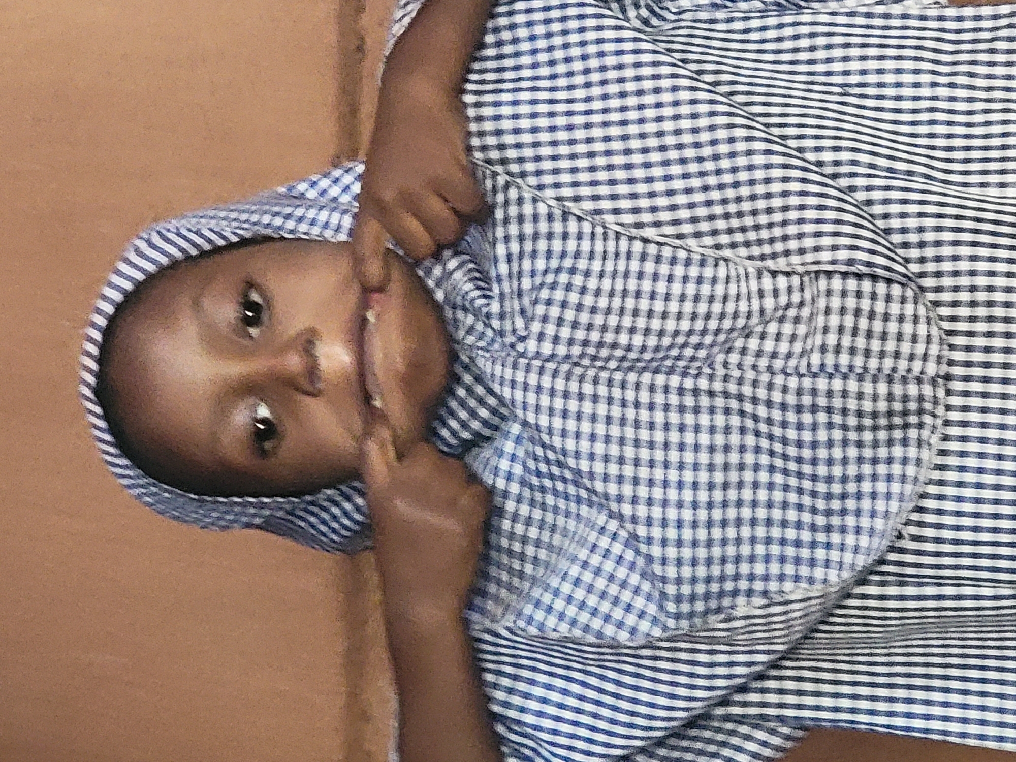 A young child with dark skin is captured in a close-up portrait against a plain, light brown background. The image is rotated 90 degrees counter-clockwise, presenting the child's head on the left side of the frame and their body extending to the right. The child is wearing a garment with two distinct blue and white patterns: a gingham check pattern is visible on the head covering and upper body, while blue and white horizontal stripes appear on other parts of the clothing, particularly on the lower visible section.
The child looks directly at the camera with wide, open eyes and a playful expression, possibly a slight smile. Both hands are raised to the child's face, with fingers touching the area around the mouth, adding to the jovial or curious demeanor. The lighting is soft and even, suggesting an indoor setting. No specific location details are discernible from the background, which is uniform in color. There is no visible text within the image. The scene conveys a sense of innocence and direct engagement from the child.