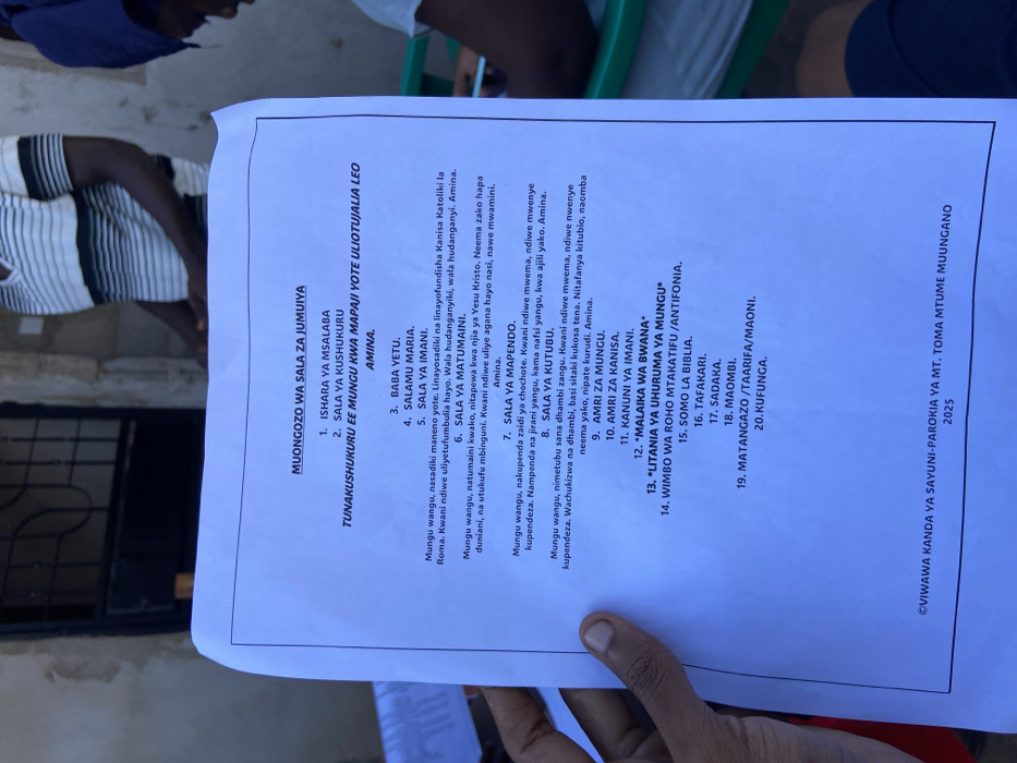 The image shows a piece of paper on a table, displaying a list of 20 Swahili phrases for prayer, titled "Muongozo wa Sala za Jumuiya" (Guide to Community Prayers). The paper is likely from a church or religious institution in Dar es Salaam, Tanzania. The location, based on the provided coordinates, is in the densely populated area of Mbezi Louis, within the Kinondoni district. The environment appears to be a simple indoor setting with a table and people in the background, though they are out of focus. The paper is the primary subject of the image, and the people in the background are not clearly identifiable. The scene is static and devoid of any particular action or interaction. The image appears to be captured with a handheld device, showcasing the paper with a slight tilt.