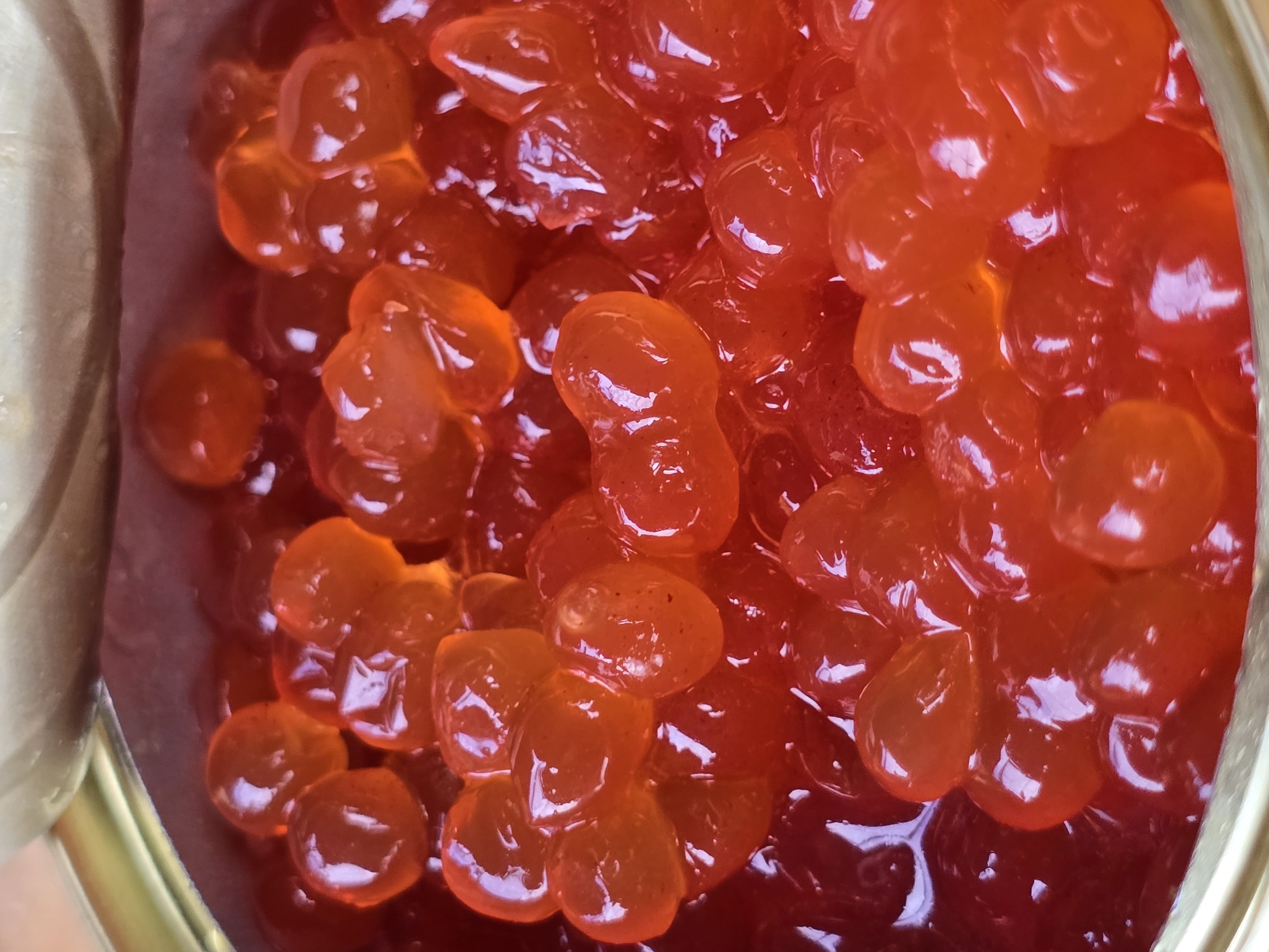 The image displays a close-up, top-down view of red caviar within a metal tin. The caviar consists of numerous small, translucent, spherical or ovular roe, with a bright orange-red hue. The roe are densely packed and appear to be glistening, reflecting the light. Some individual roe show slight indentations or irregularities in shape. The tin's rim, a metallic gold color, is partially visible at the bottom and along the left edge. The background consists of the interior of the tin, which also appears to be metal. There are no people, animals, or other objects present. The scene is domestic or food-related. The provided location information is irrelevant to the visual content of the image.