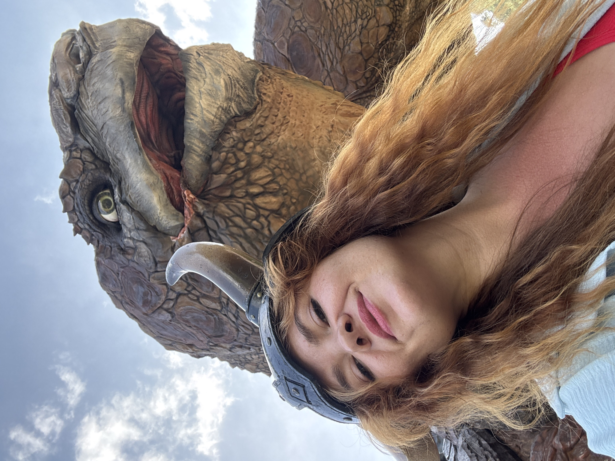 An outdoor, ground-level, close-up selfie shows a smiling young woman with long, wavy, red-blonde hair. She is wearing a black helmet with a horn and a light blue shirt over a red garment. Behind her, a large, reptilian creature, possibly a dinosaur or dragon, with scaly, brown skin and a wide-open mouth showing red interior and sharp teeth, is partially visible against a cloudy blue sky. The scene appears to be in an amusement park or a themed attraction.