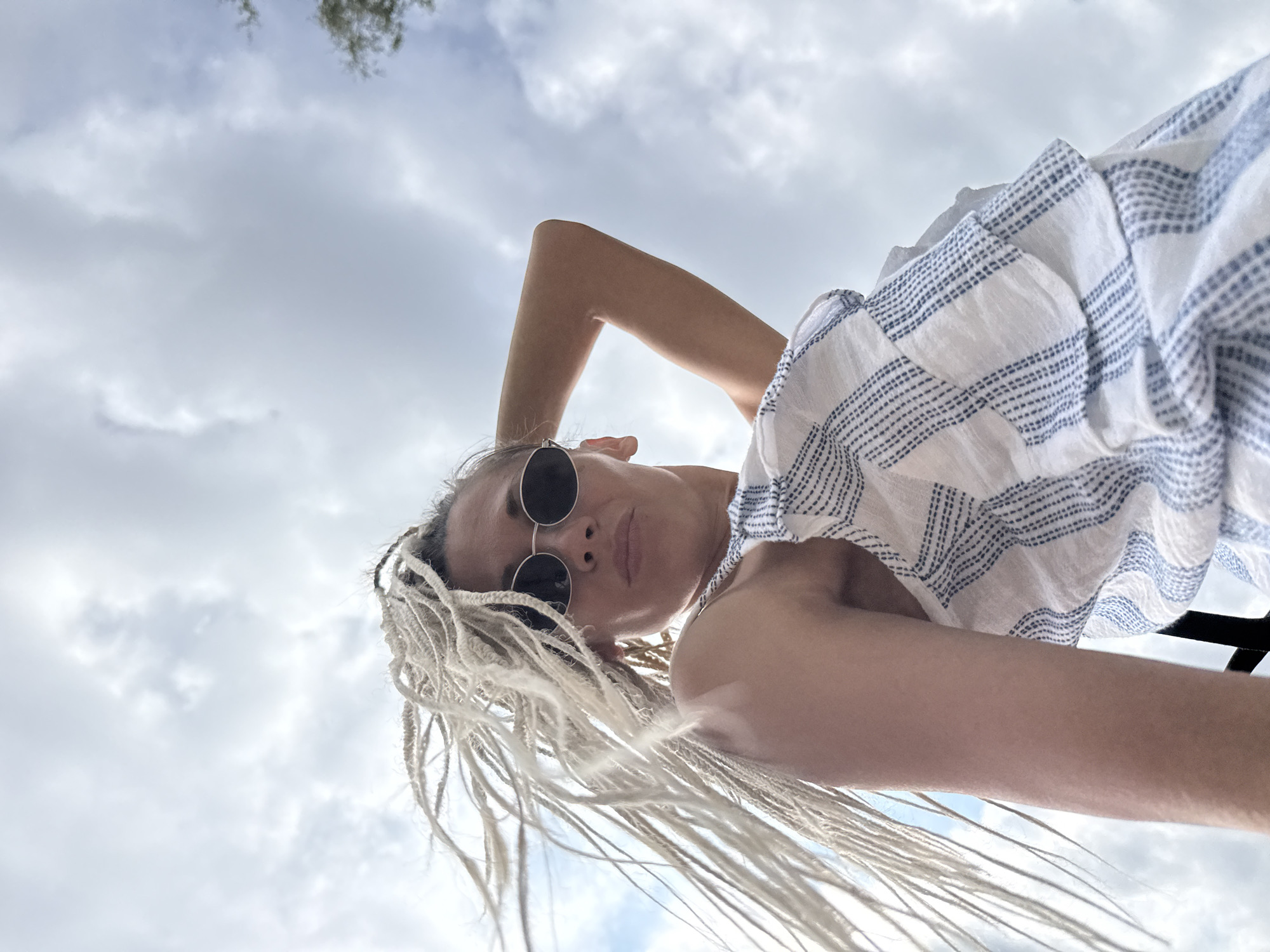An outdoor, low-angle, medium shot captures a woman with light blonde braids against a cloudy sky. The woman is wearing round, silver-rimmed sunglasses and a white top with blue stripes and ruffles. Her right arm is bent at the elbow, with her hand resting behind her head. Her braids are spread out, moving to the left due to wind. The scene is set in the city of Rusanovo, Russia.