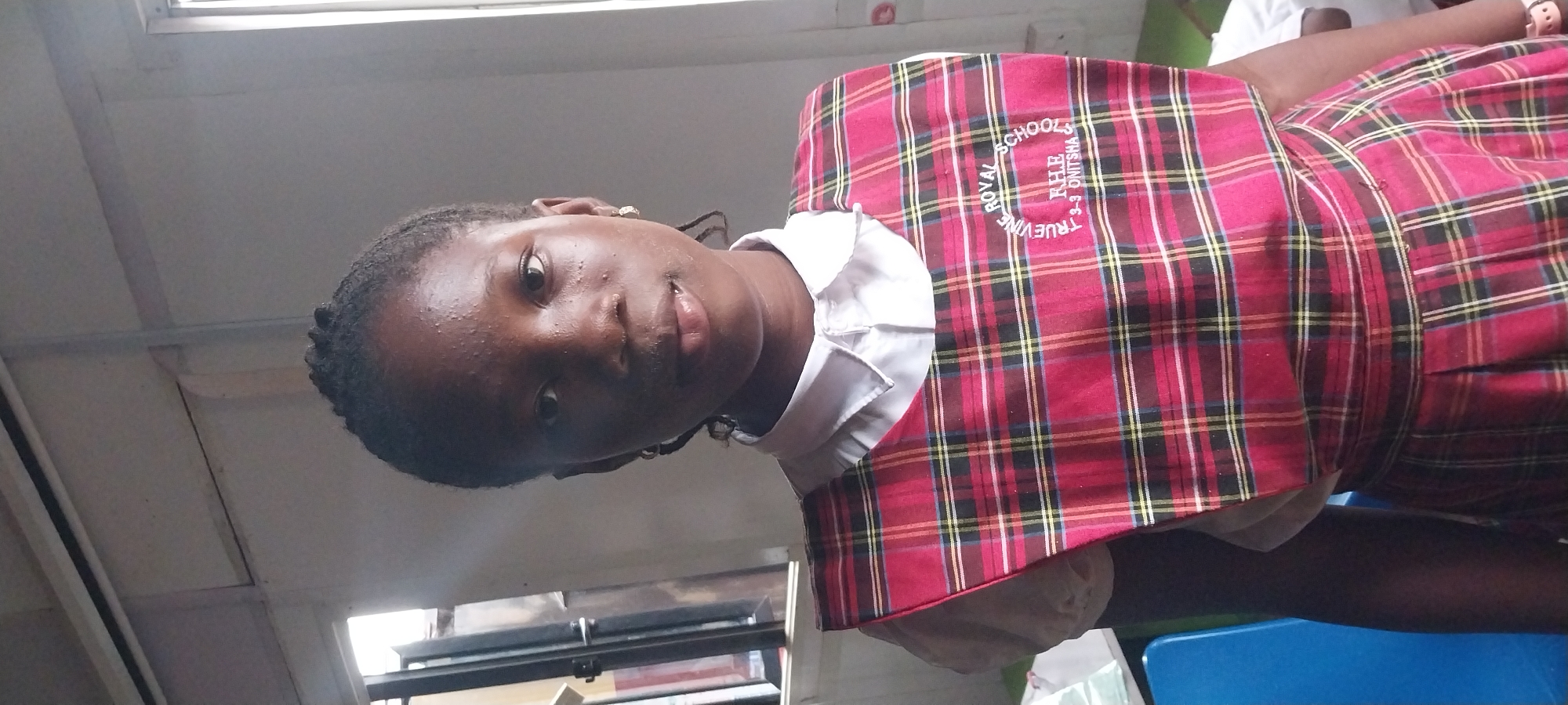 A young Black girl, likely of school age, is depicted from the chest up in an indoor setting. She is wearing a white collared shirt beneath a red, plaid jumper dress. The dress features a circular embroidered emblem on the chest that reads "TRUEVINE ROYAL SCHOOLS" with "RHE 3-3-3 ONITSHA" below it. Her hair is styled in braids, and she has small earrings in her ears. The background suggests a classroom or similar educational environment, with a window visible on the left side of the frame and a hint of blue furniture on the far left. The lighting is somewhat harsh, casting shadows and highlighting some shine on her skin. The overall impression is a posed, head-and-shoulders portrait taken within the Truevine Royal Schools in Onitsha, Nigeria.