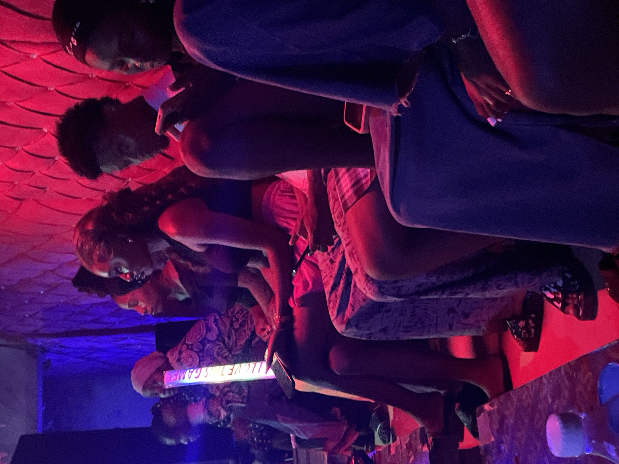 The image depicts a group of individuals, primarily women, seated in a dimly lit environment with vibrant red and blue lighting. They appear to be in a social setting, possibly a club or lounge. The individuals are positioned closely together, suggesting a shared activity or conversation. One person is holding a glowing sign that reads "I LOVE THIS GAME". Another person is looking at a mobile phone. The clothing styles suggest a contemporary setting. The environment is characterized by textured walls, likely upholstered, and a floor that reflects the colored lighting. The overall ambiance is festive and possibly energetic. The specific location in Durumi, Nigeria, is not explicitly indicated by any visual cues in the image.