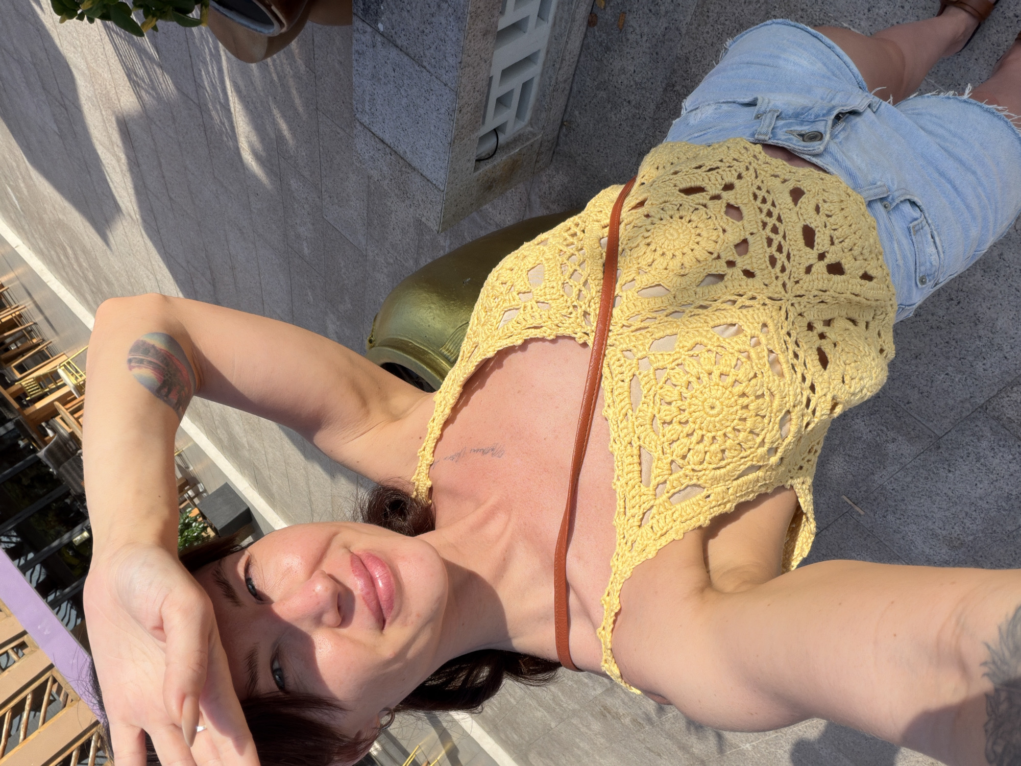 In this selfie taken outdoors, a woman with fair skin and dark hair pulled back is shielding her eyes from the sun with her right hand, which is placed on her forehead. She is wearing a yellow crocheted halter top and light blue denim shorts. A brown strap from a bag is visible across her chest. There is a tattoo on her upper left arm. The background shows a paved area with shadows cast by what appears to be outdoor seating and structures, suggesting a public space like a cafe or restaurant. The environment is brightly lit by sunlight, indicating a daytime setting. The location is identified as Dong Ninh Hoa Ward, Vietnam, though specific landmarks to confirm this are not clearly visible in the image itself.