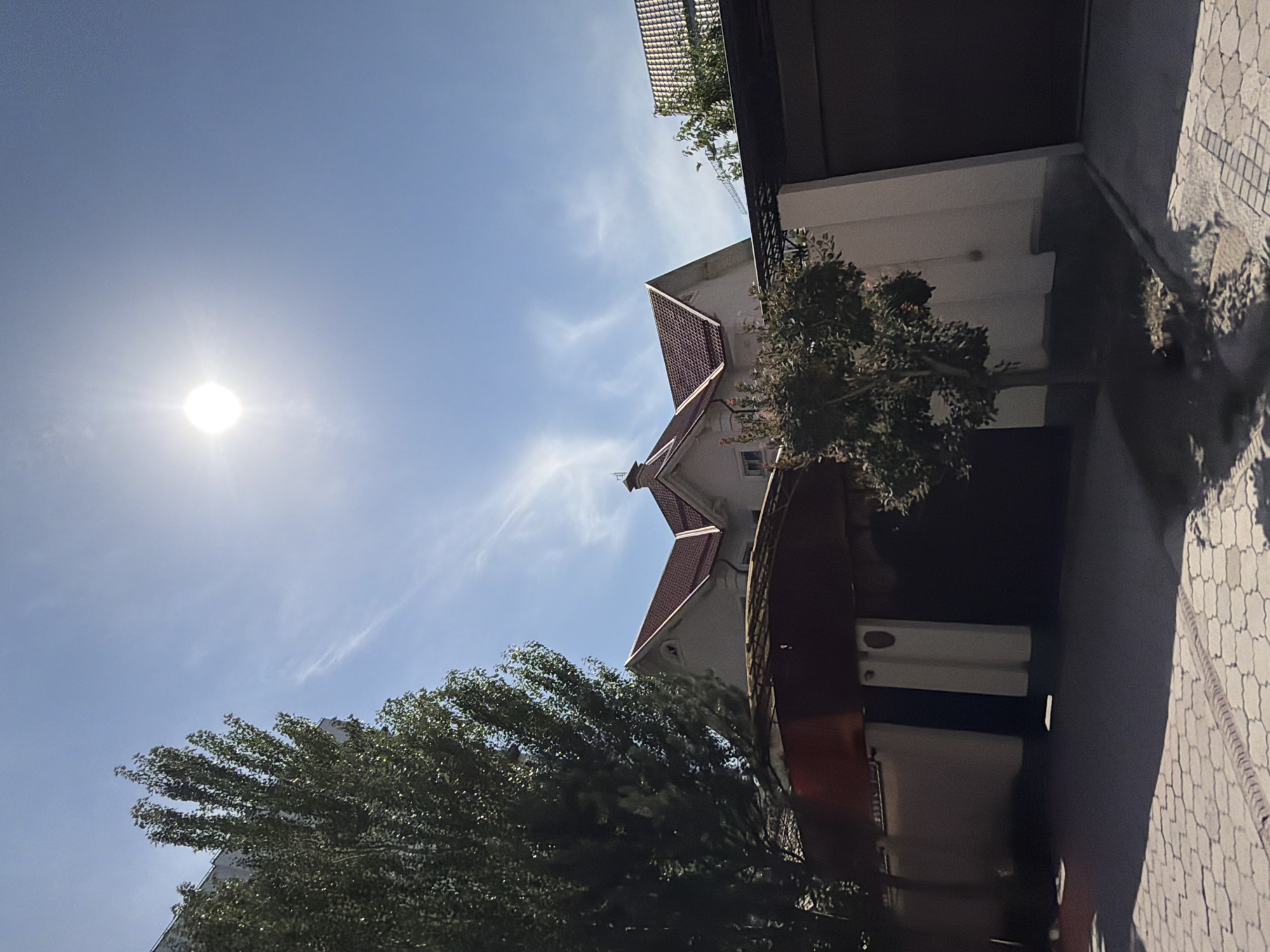 A low-angle shot captures a bright sun in a clear blue sky, with thin wisps of clouds. The sun is positioned in the upper center of the frame. In the foreground, a tall, leafy green tree with dark bark stands to the left. To the right of this tree, a portion of a building with a reddish-brown tiled roof is visible. The building appears to be a house with light-colored walls. A dark, enclosed space, possibly a garage or gate, occupies the right side of the frame. The ground in the foreground is paved with light-colored interlocking stones.