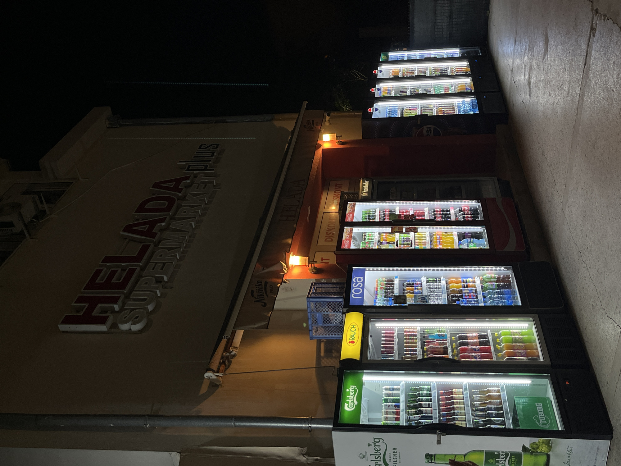 The image depicts a night scene outside a "HELADA SUPERMARKET plus" in Budva, Montenegro. The supermarket facade features illuminated signage. In front of the supermarket, several glass-fronted refrigerators are lined up, stocked with various beverages. The refrigerators display prominent branding such as "Carlsberg", "RAUCH", and "rosa". Additional refrigerated units further down the line show "Coca-Cola" and "Pepsi" branding. The ground is paved with concrete, showing some cracks and wear. A vertical pipe runs along the left side of the building. The lighting is primarily from the illuminated signs and the interior lights of the refrigerators, casting a glow on the surrounding pavement and building. There are no people visible in the image.