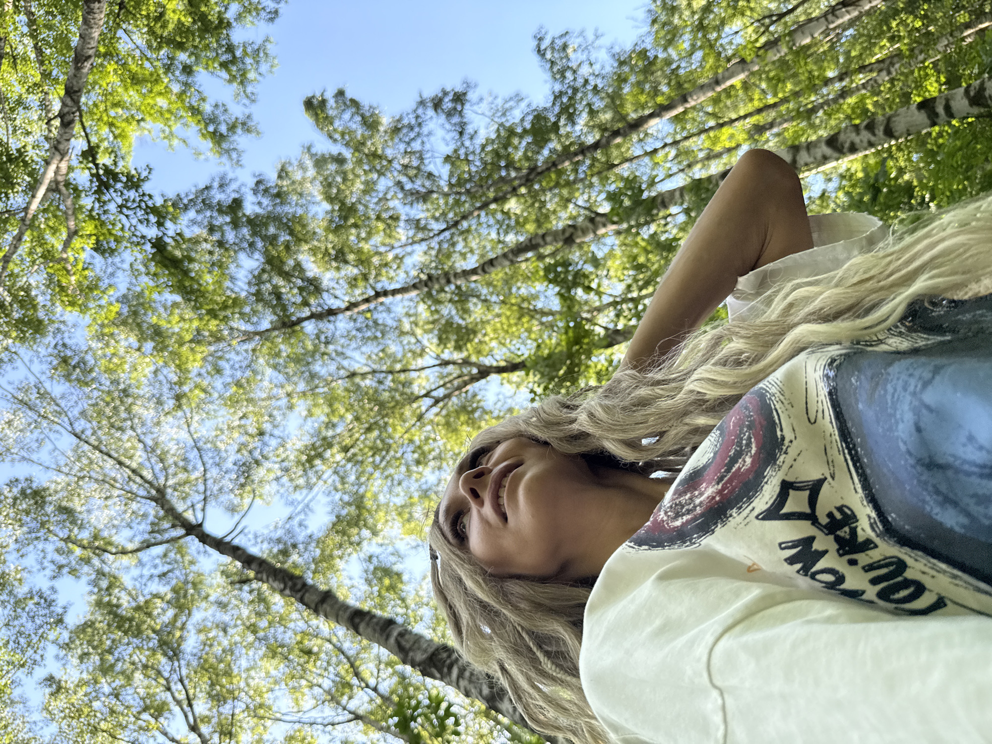 An outdoor, low-angle, medium shot captures a Caucasian woman with long, blonde, braided hair. She is wearing a white t-shirt with a graphic design. Her face is turned upwards and to the left, with her eyes looking in that direction and her mouth slightly open. Her right arm is bent, with her hand resting near her head, partially obscuring her hair. The background consists of tall birch trees with green foliage, against a clear blue sky, suggesting a forest or park setting in Rusanovo, Russia. The lighting is natural and bright, indicating daytime.