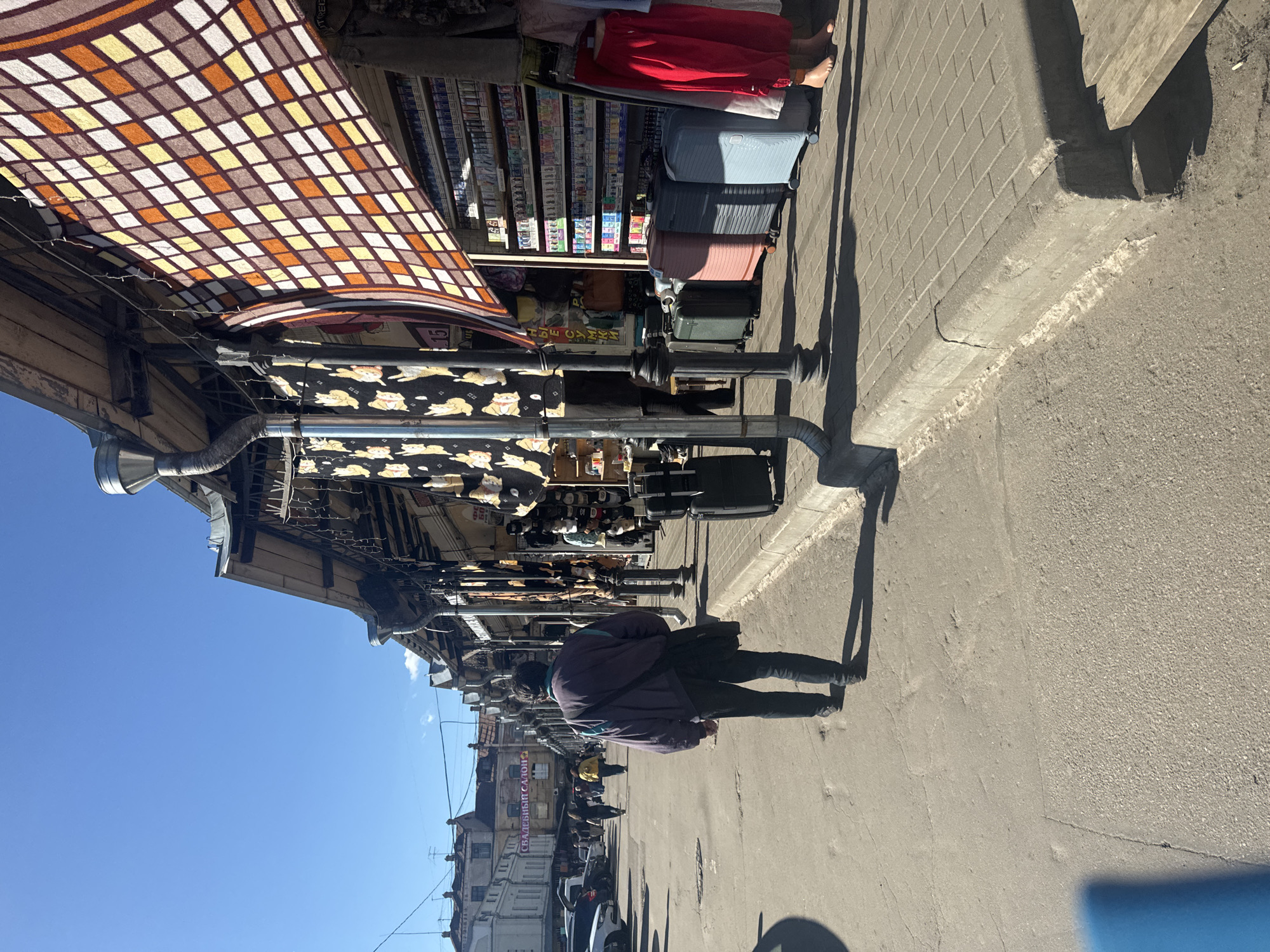 This image depicts an outdoor market scene, likely in Saint Petersburg, Russia, under a clear blue sky, suggesting a sunny day. The perspective is from a low angle, looking upwards towards a covered walkway lined with stalls. A person, seen from behind, walks away from the viewer on a paved sidewalk. They are wearing a dark jacket and pants, with a bag slung over their shoulder.
The stalls feature various goods, including textiles with a checkered pattern draped overhead, and what appears to be a display of suitcases stacked against a building. Further down the row, shelves are filled with items, possibly cigarettes or small packaged goods. Another stall displays fabric with a pattern of cartoonish foxes.
In the background, buildings with multiple windows and architectural details are visible, hinting at an urban environment. One building has Cyrillic text that reads "СВАДЕБНЫЙ САЛОН" which translates to "Wedding Salon." The overall impression is one of a bustling marketplace on a bright day, with the interplay of sunlight and shadow casting long shapes on the pavement. The scene appears to be active, with indications of commerce and pedestrian traffic.
