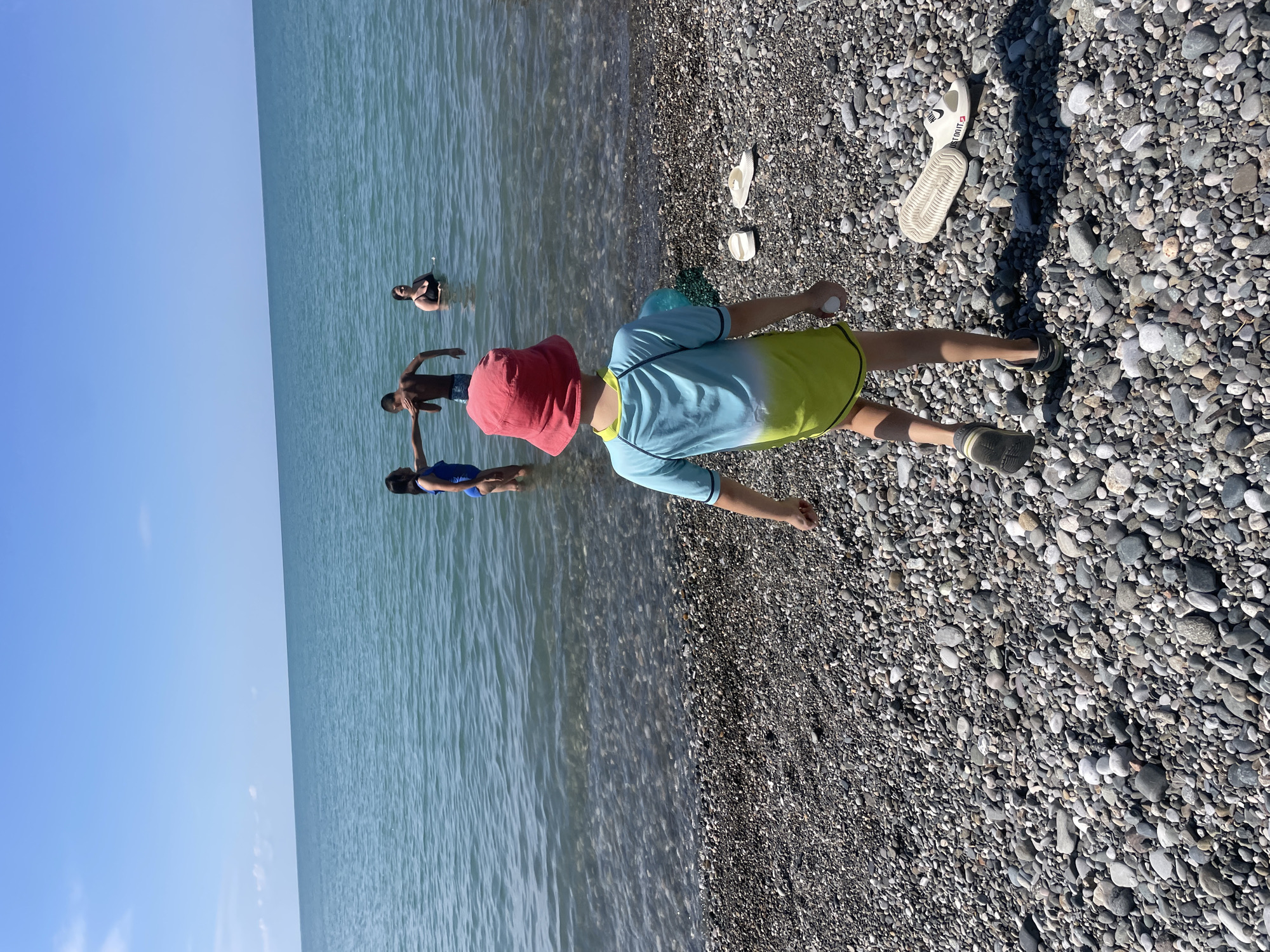 In Giachrypsh, Abkhazia, a sunny day unfolds at a pebble beach. The turquoise sea, calm and clear, stretches to the horizon under a bright blue sky. In the shallow water, four individuals are present. A woman in a blue swimsuit stands on the left, facing right. Next to her, a shirtless boy in dark swim trunks faces left, his arm extended as if interacting with the woman. Further back, a woman in a black swimsuit is submerged up to her chest. Another individual, barely visible, is in the water behind the boy.
On the shore in the foreground, a child wearing a red bucket hat and a blue and yellow swimsuit faces away from the camera, walking towards the sea. The child holds a small white object in their right hand. To the child's right, two pairs of white flip-flops lie on the pebble-strewn beach. The pebbles are a mix of gray, brown, and white, smoothed by the water. The overall scene depicts a relaxed summer day at the seaside.