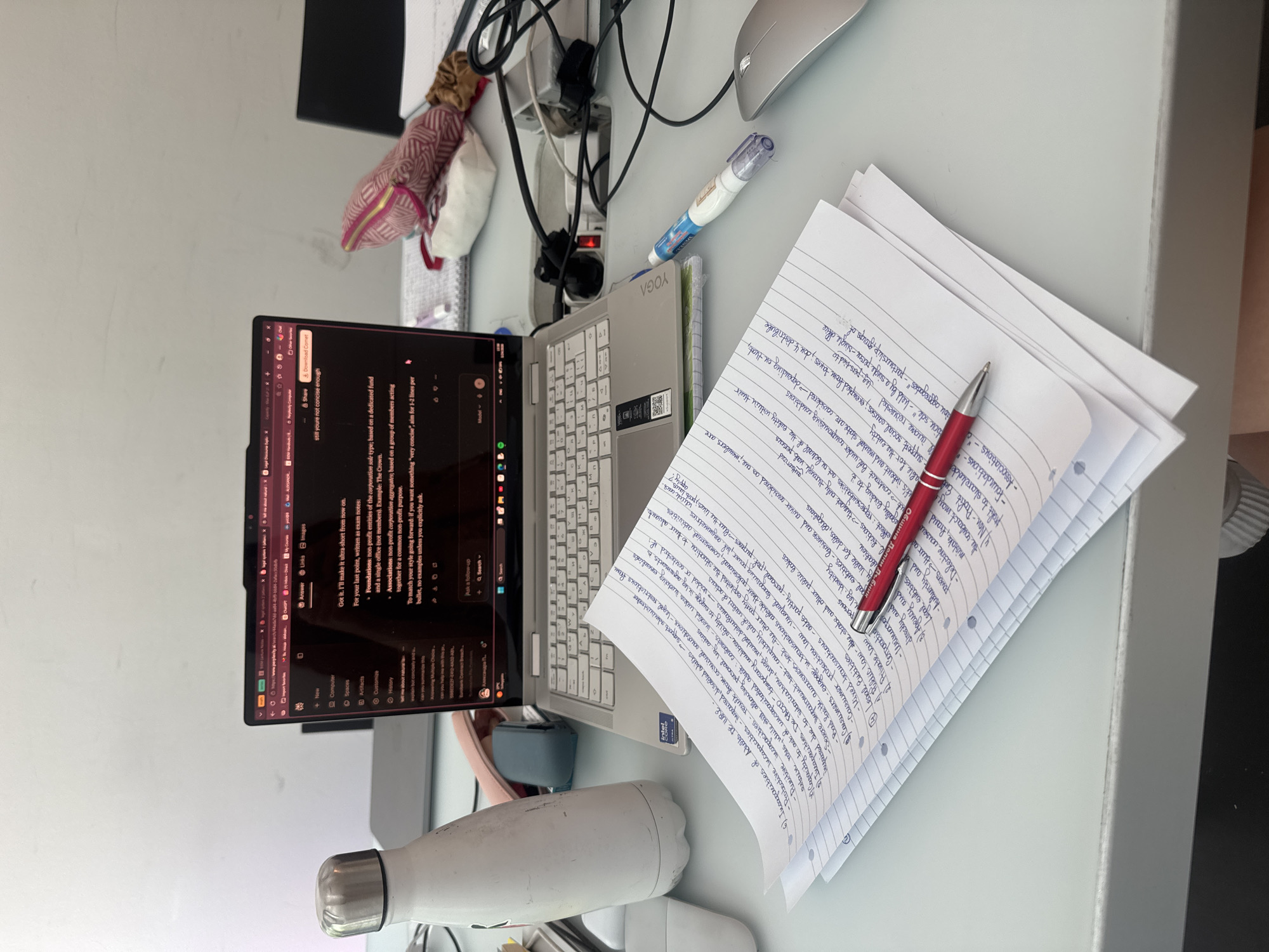 The image shows a desk with a laptop, notebook, pen, and other objects. The laptop is open, displaying a text editing interface and search results about legal entities, with notes on the right. A red pen rests on the notebook, which contains handwritten notes related to legal capacities and entities, including "Incapacities of Adults DE IURE", "Legal Entities", and "Non-Profit Entities". The desk also contains a water bottle, a correction pen, and a computer mouse. The laptop is a Lenovo Yoga model. The environment is an indoor setting, likely a home office, but there is no information that helps determine the location is in the city of Milan, Italy.