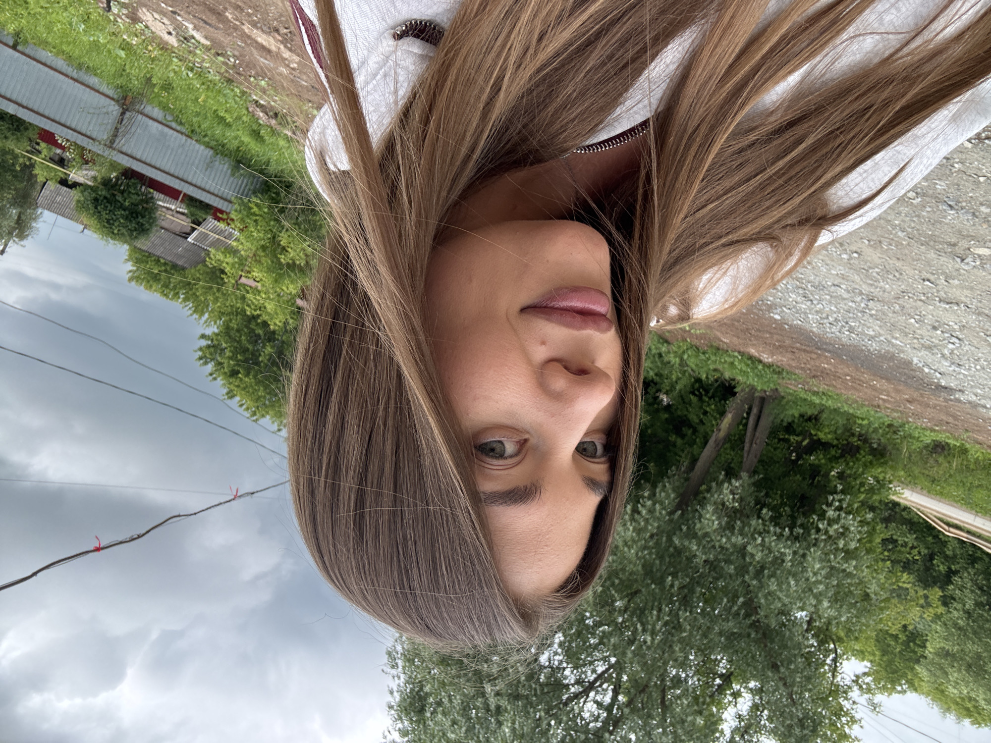 A young woman with long, light brown hair takes a selfie outdoors in Chernushka, Russia. She is wearing a white hoodie. The background includes trees, a dirt road, and a grey, overcast sky. In the distance, there are houses and a metal fence.