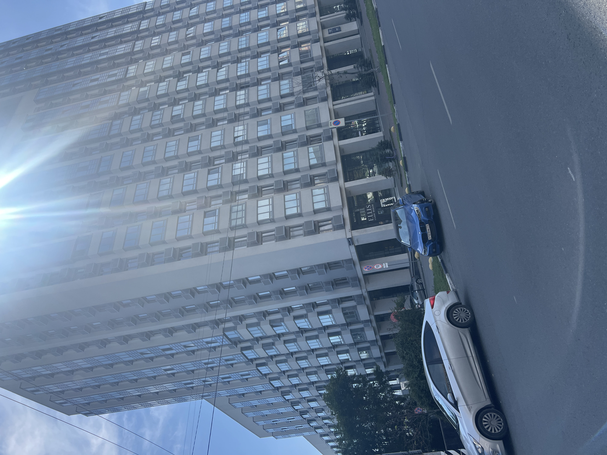This is a photograph taken from a street level perspective, capturing a section of a modern, tall apartment building. The building is light gray and features a uniform design with numerous windows and balconies. The sun is shining brightly, creating a lens flare effect that partially obscures the top of the building and the sky. In the foreground, there are two cars parked on the side of the road: a silver sedan and a blue sports car. The street is paved, and there is a sidewalk and some greenery along the side of the building. The image appears to have been taken in an urban environment, likely a residential area. Based on the license plates visible on the cars, specifically the blue car with "P 123 MK 73" and the silver car with "H 785 AE 73", the location is identified as Ulyanovsk, Russia, as the "73" region code indicates.