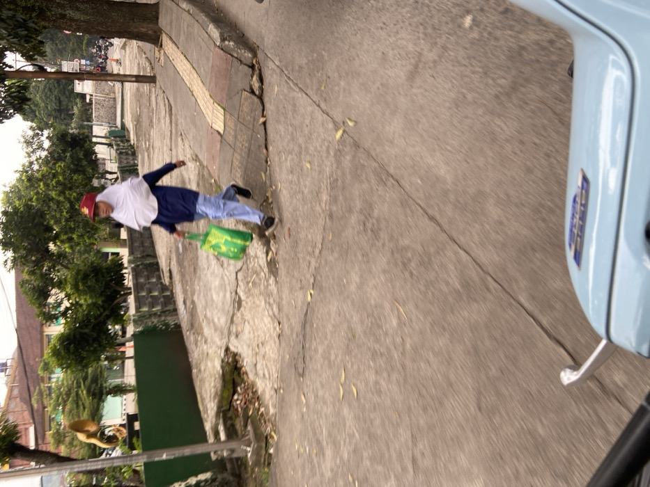 The image shows a person walking on a sidewalk in the city of Bandung, Indonesia. The person is wearing a white hijab and a blue shirt. The person is carrying a green bag and is walking towards the right. The image was taken from a moving vehicle, likely a motorcycle, which is visible in the bottom of the frame. The sidewalk is cracked and uneven. There are trees and bushes lining the sidewalk. The location is in the city of Bandung, Indonesia, based on the coordinates provided. The coordinates suggest that the image was taken in a residential or commercial area.