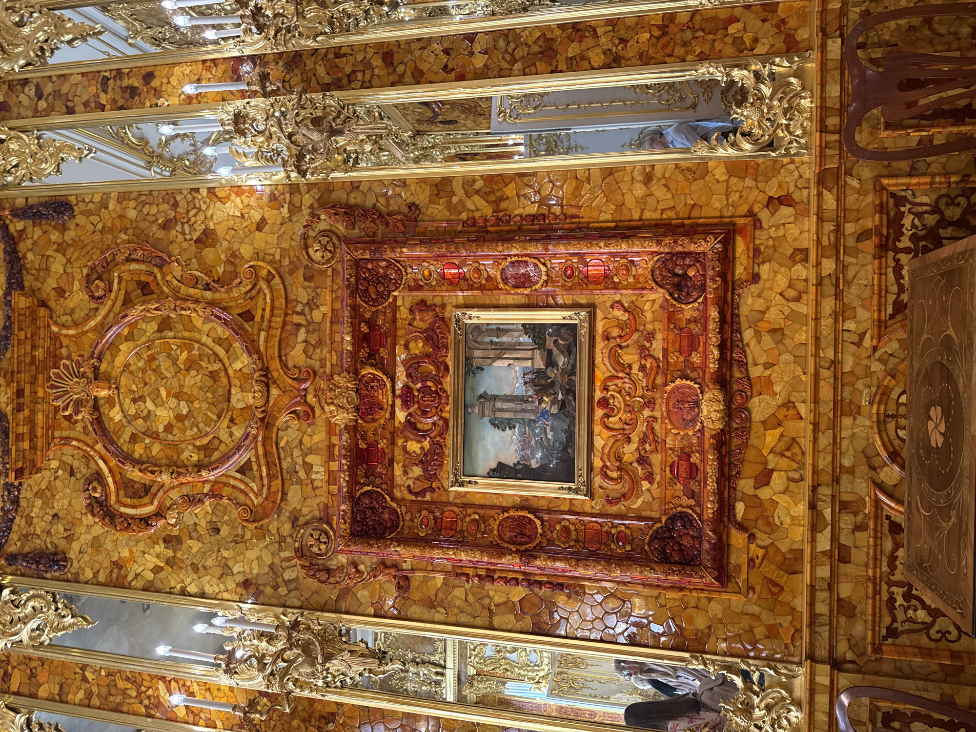 The image shows a close-up of a wall in the Amber Room, located in the Catherine Palace in Pushkin, Russia. The wall is extensively decorated with pieces of amber, creating a warm, honey-like hue. Intricate gilded frames surround a central framed painting, which depicts a classical landscape scene with figures. The frames themselves are also ornately carved and decorated with what appears to be amber or gilded wood elements. To the sides of the central artwork, tall, slender mirrors framed in elaborate gold leaf reflect the opulent surroundings and people visiting the room. Several candelabra with electric candles are attached to the gold frames on the left side. In the foreground, the top of a wooden table with inlay work and part of a wooden chair are visible, indicating the presence of furniture within the room. People are partially visible in the reflections of the mirrors, suggesting the space is open to visitors. The overall impression is one of immense wealth and detailed craftsmanship.