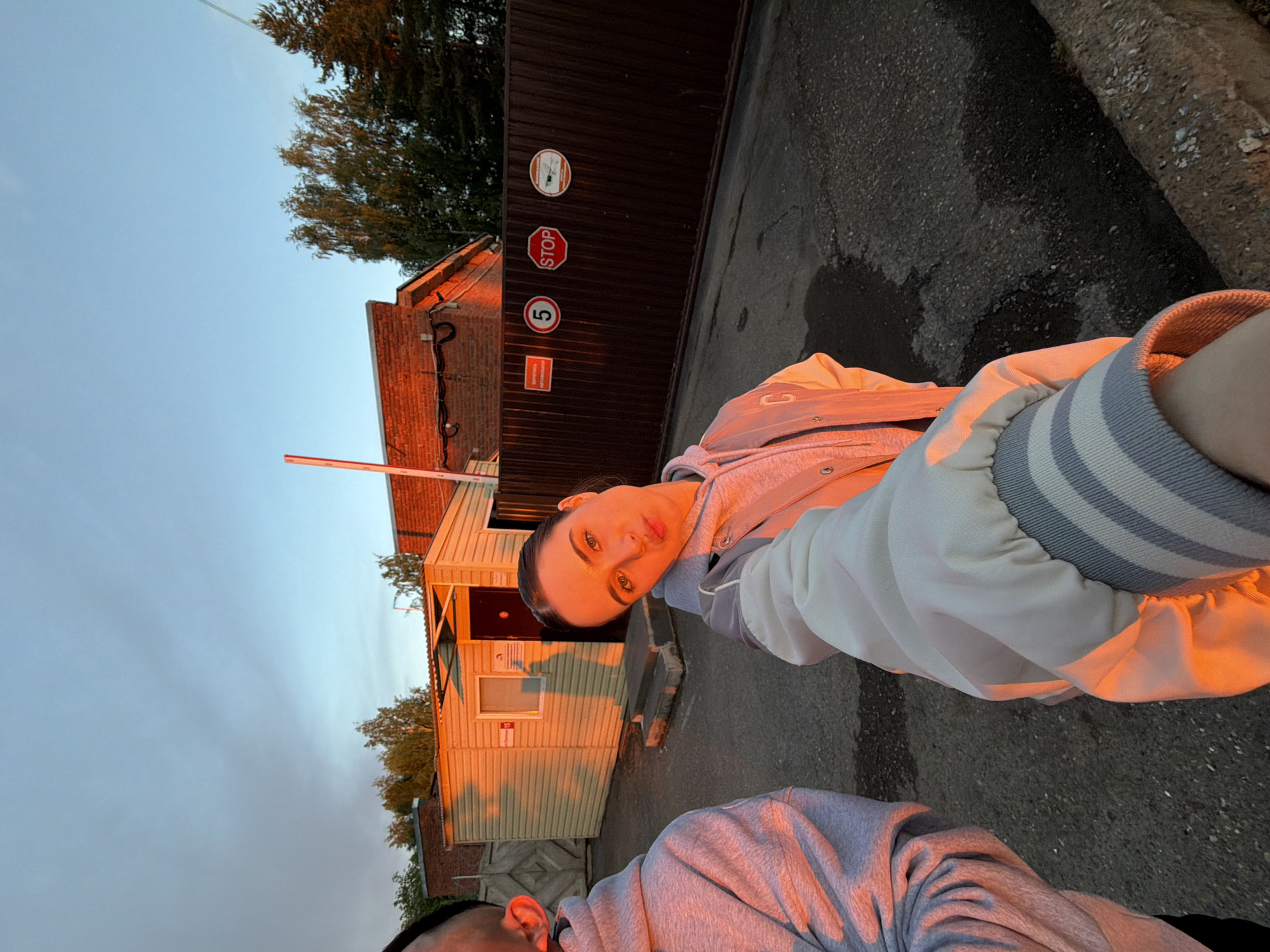 A selfie photograph captures two individuals in what appears to be Izhevsk, Russia. The woman, positioned closer to the camera, has dark hair pulled back and fair skin. She gazes directly into the camera. The other person is cut off at the bottom of the frame, but is facing the woman. Both individuals are wearing matching grey hoodies.
Behind the subjects, a small, light-colored building with a closed door and window is visible. Adjacent to this is a brick structure, and a dark metal gate with a "STOP" sign and speed limit indicator on it. The ground is paved with asphalt. The sky is overcast, suggesting it is either early morning or late evening. Golden hour lighting suggests the sun is either rising or setting.