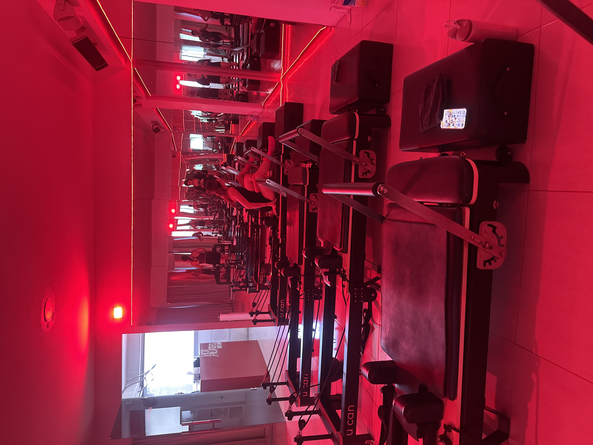 The image shows the interior of a brightly lit studio. The room is filled with Pilates reformer machines, arranged in rows. Each reformer has a sliding carriage, springs, and foot bar. In the background, a large mirror reflects the rows of reformers and the red lighting. The red lighting illuminates the room, casting a strong hue over everything. Some of the machines are in use by people who are wearing black clothing. The window in the background shows some signs for 'HOT PILATES', and 'U CAN'. The signage indicates that the location is most likely in the city of Puebla, Mexico.