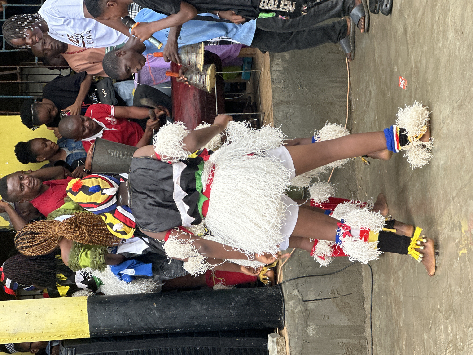 This is a medium shot, taken from a low angle, of a group of people gathered outdoors, likely in Badagry, Nigeria. The foreground features two individuals dressed in traditional attire for a performance. The person on the left is mostly out of frame, but their legs are visible, adorned with white fringed leg warmers and yellow ankle bells. The central figure, facing away from the camera, wears a black and white top with colorful trim, a white fringed skirt, and elaborate headwear. Their legs are similarly decorated with white fringe and colorful bands around the calves.
Behind these performers, a crowd of onlookers watches. To the right, a person in a blue shirt holds two bell-like instruments, possibly playing them. Another person in the background wears a red shirt. The environment appears to be an outdoor public space with a concrete ground. A yellow and black structure, possibly a wall or pillar, is visible on the left, along with a large black speaker. The overall atmosphere suggests a lively cultural event or performance.