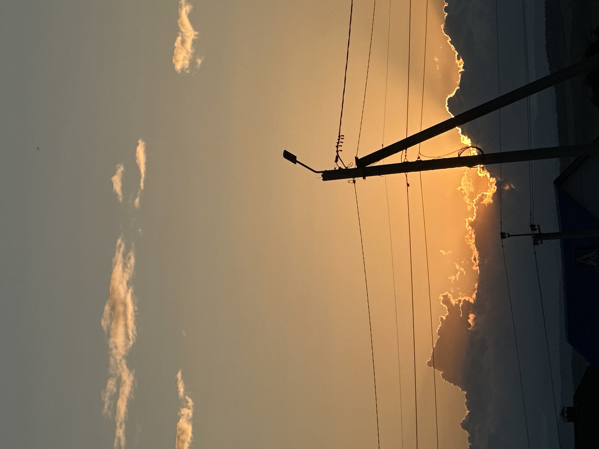 This media file depicts a sky at sunset, with a prominent utility pole silhouetted against the warm hues of the sky. The sun is setting behind a line of clouds, creating a dramatic contrast between the bright, illuminated edges of the clouds and the darker, shadowed portions. Thin, wispy clouds are scattered across the left side of the sky, which is a lighter shade of orange and yellow. The utility pole, made of dark wood, has several wires running from it, also silhouetted against the sky. One of the wires extends horizontally across the frame, and another slanted wire connects to a street lamp. The overall mood is serene and slightly dramatic due to the strong contrast of light and shadow. The time of day is clearly sunset, indicated by the warm, golden light. There is no visible text in the image.