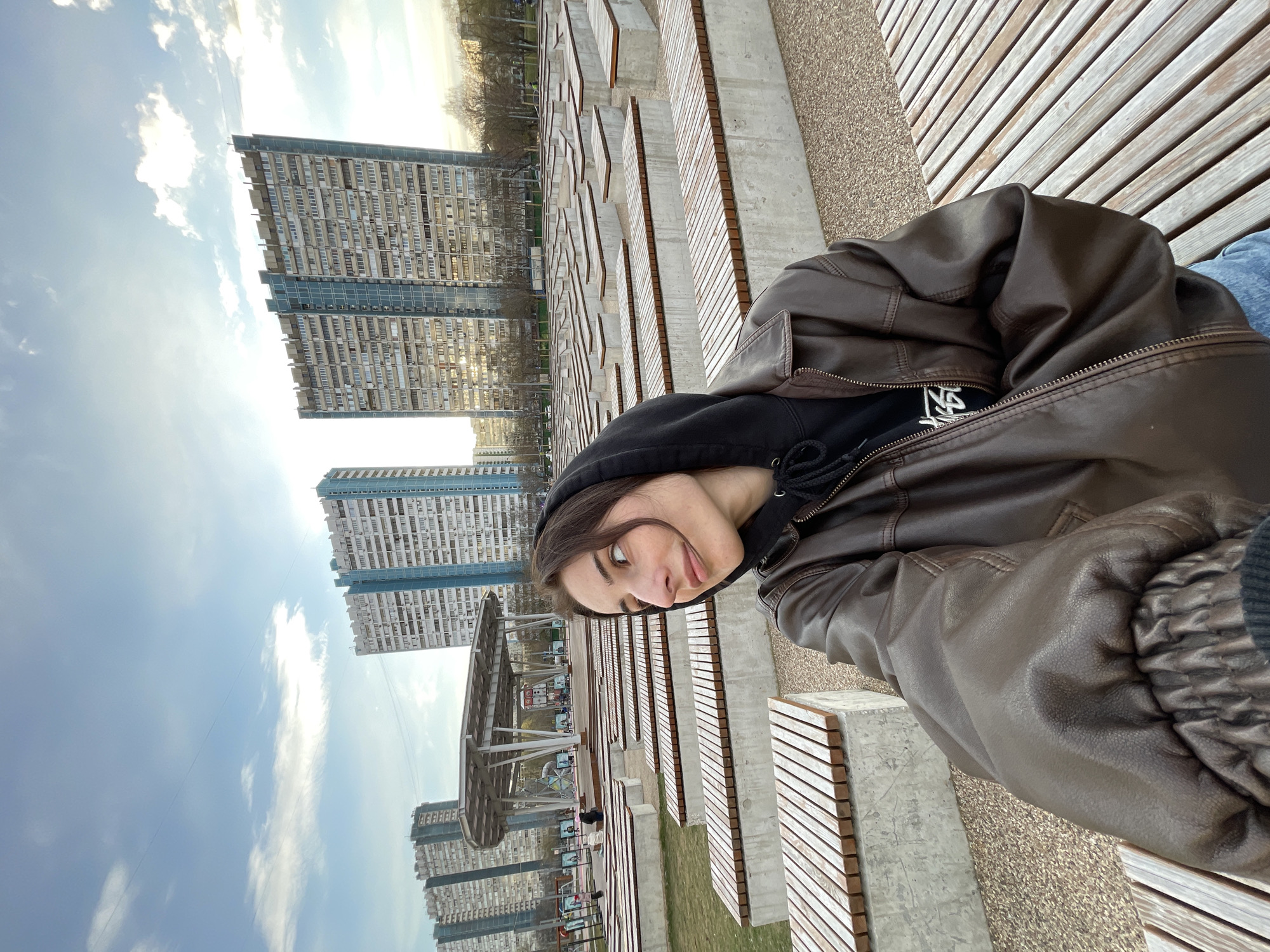 A vertically-oriented, medium shot shows a person sitting on a wooden bench in a public space with modern architecture. The person is wearing a black hooded sweatshirt and a brown leather jacket. In the background, there are several tall apartment buildings with a repetitive window pattern, typical of Soviet-era architecture, which suggests the location may be in a residential area of Moscow, Russia. The sky is partly cloudy with the sun visible in the upper left of the image. There are multiple rows of benches with concrete supports, creating a layered, geometric pattern extending into the background. There is what appears to be a modern park or gathering space with metal framework structures and possible recreational equipment between the benches and the apartment buildings. The person is looking at the camera and appears to be taking a selfie.