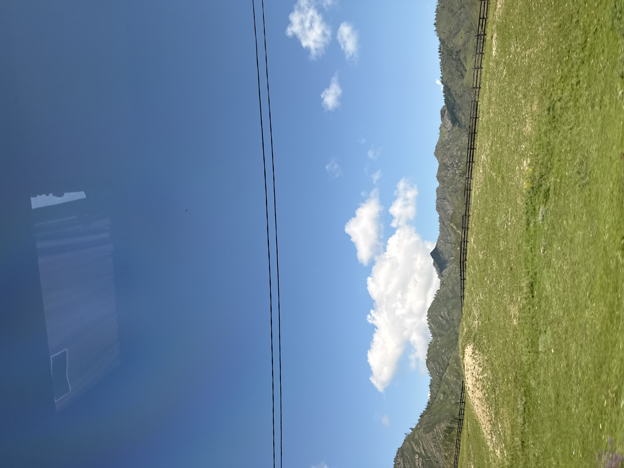 The image shows a pastoral landscape under a bright blue sky with scattered white clouds. In the foreground, a vibrant green, grassy hill slopes upwards, with a wooden fence running horizontally across its middle. Beyond the hill, a range of green, tree-covered mountains forms the horizon. Two dark electrical wires are stretched across the upper portion of the sky. In the top section of the image, a reflection is visible on what appears to be a window, showing a white, rectangular shape and vertical lines. The scene is indicative of the natural beauty found in rural areas like Tuekta, Russia.