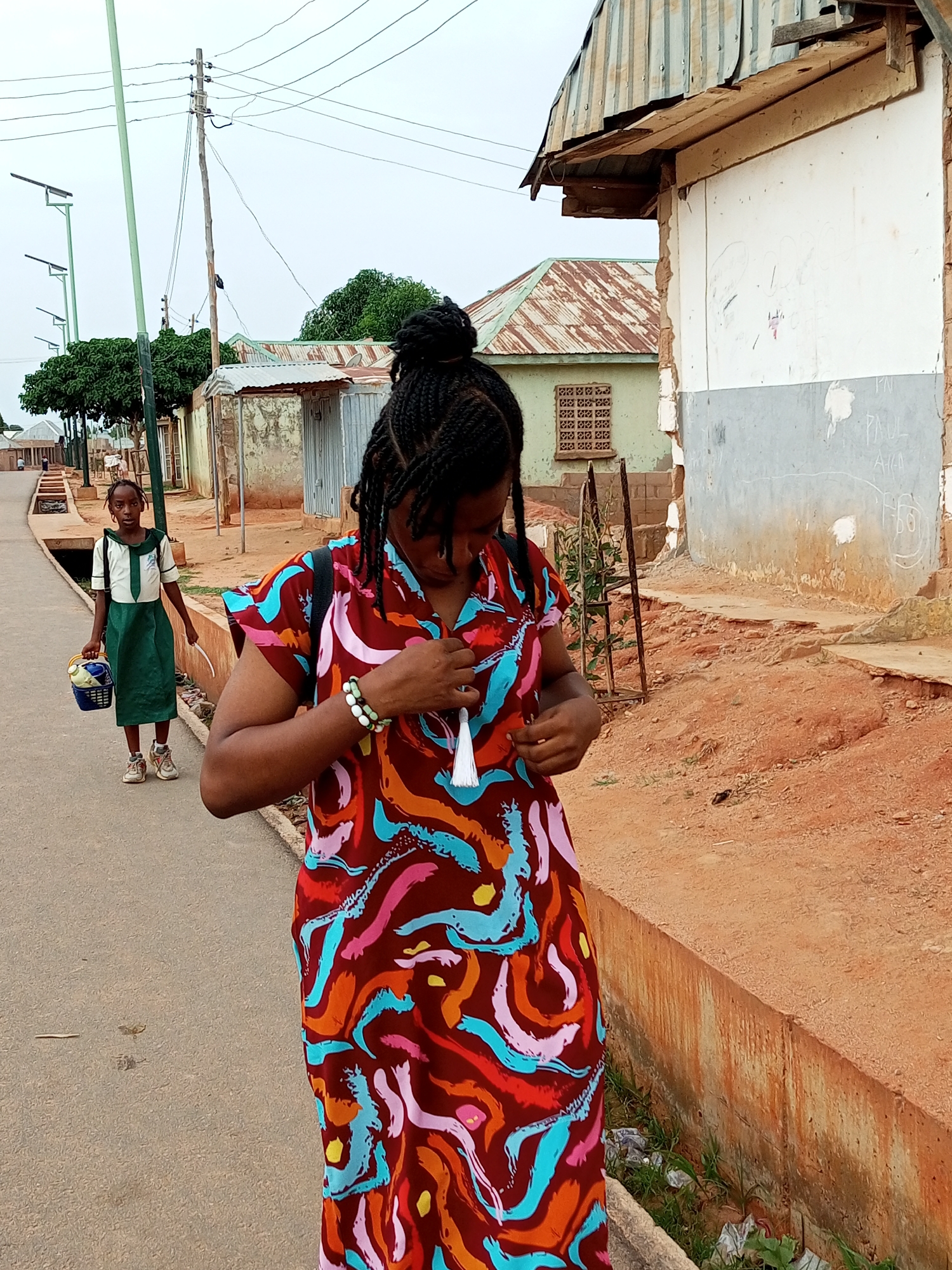 In Tempure, Nigeria, a young girl in a school uniform walks on the left side of a paved road, carrying a basket and a white object. A woman in a colorful dress stands on the right, adjusting her attire. The scene unfolds in a dusty, urban environment characterized by unfinished buildings with corrugated metal roofs, utility poles with overhead wires, and sparse vegetation. The road is flanked by a concrete drain on the right, filled with debris. The sky is overcast, suggesting a cloudy day.