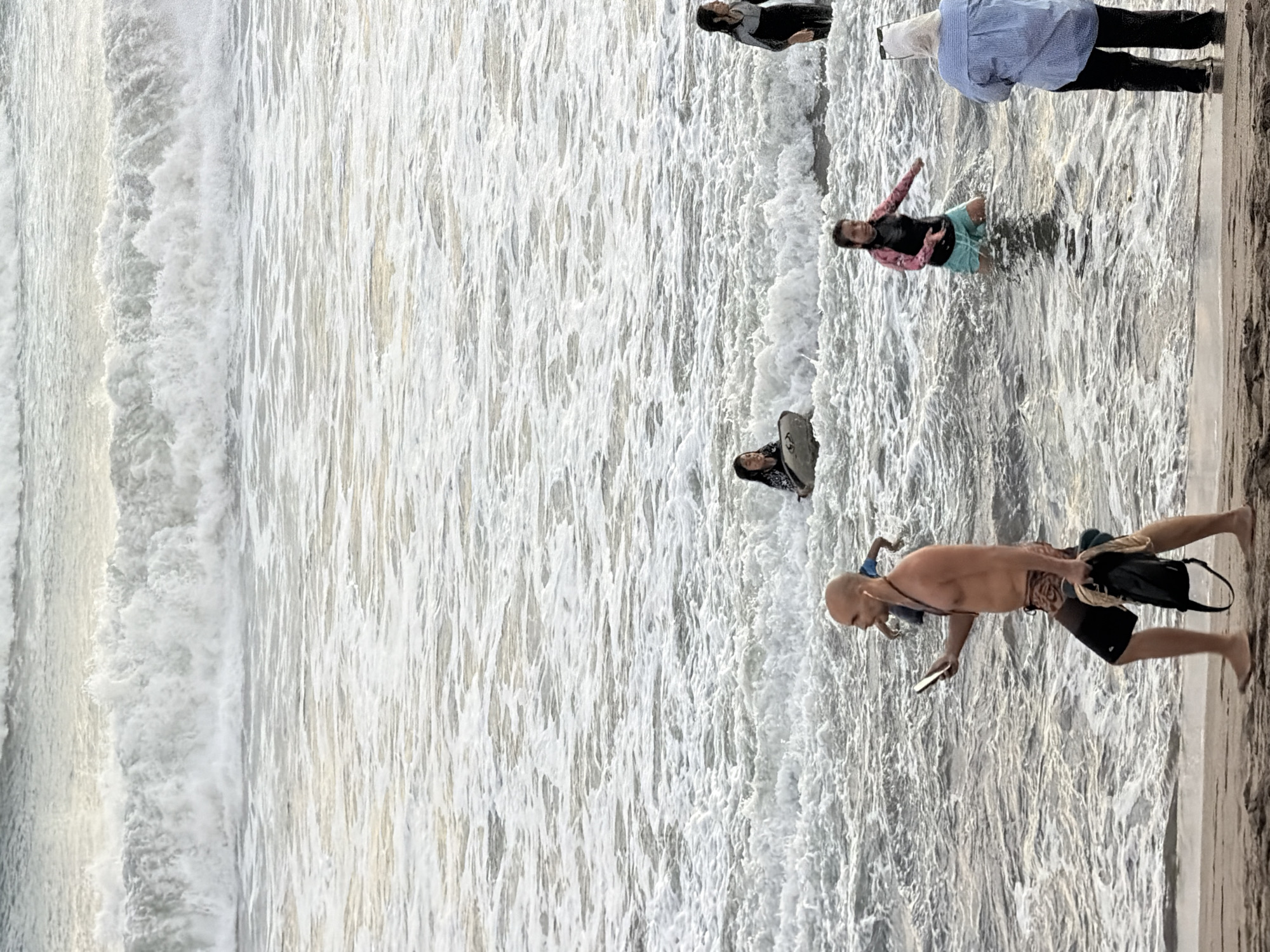 In North Kuta, Indonesia, a beach scene unfolds with several individuals in the shallow ocean and on the shoreline. In the foreground, a bald man in patterned swim trunks walks on the sand, holding a smartphone in his left hand and a dark bag with rope in his right. He is wearing earbuds. Further out in the water, a young girl in a pink and black rash guard and turquoise shorts kneels, looking towards the left. To her right, a woman wearing a blue striped shirt and black pants stands on the sand, facing the ocean. In the mid-ground, a young girl in a dark patterned rash guard sits on a surfboard, while another young girl in a dark full-body swimsuit stands to her right, also in the water. The ocean is choppy with significant wave activity and foam. In the background, large waves crash. The overall lighting suggests it is either early morning or late afternoon.