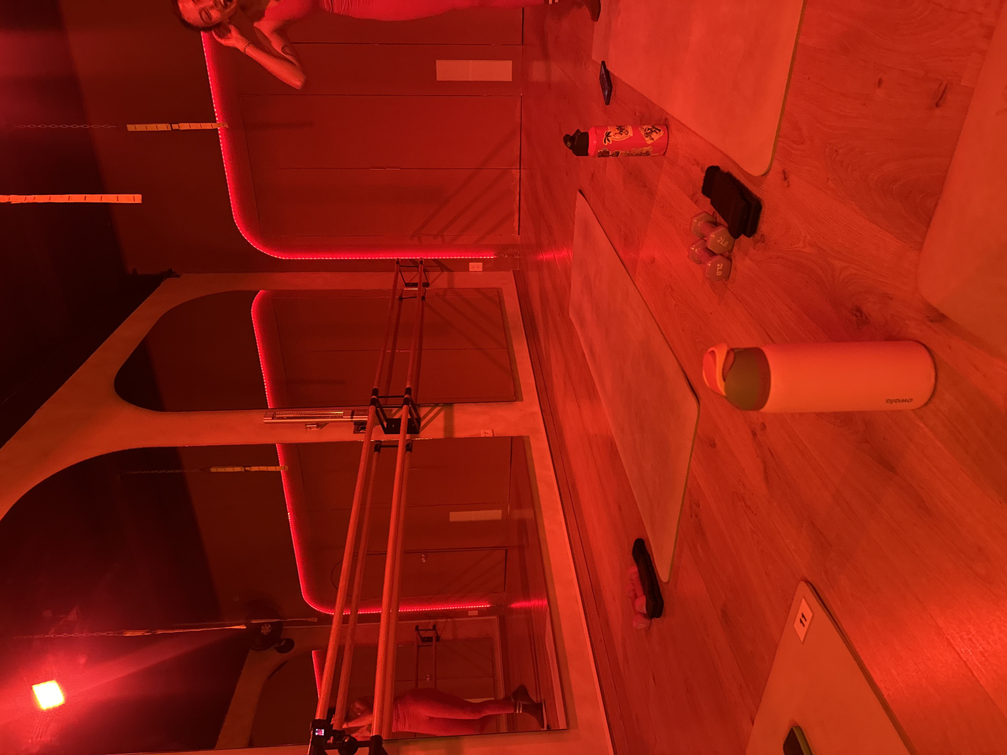The image shows a room interior, possibly a fitness studio, illuminated by strong red light. In the reflection of a mirror, a person can be seen lying on a surface. Two exercise mats are present, along with various objects including weights, a water bottle labeled "owala," and a phone. A structure with wooden poles and metal bars is also visible. The number "11" is seen on a mat. The specific location is not evident from the image.