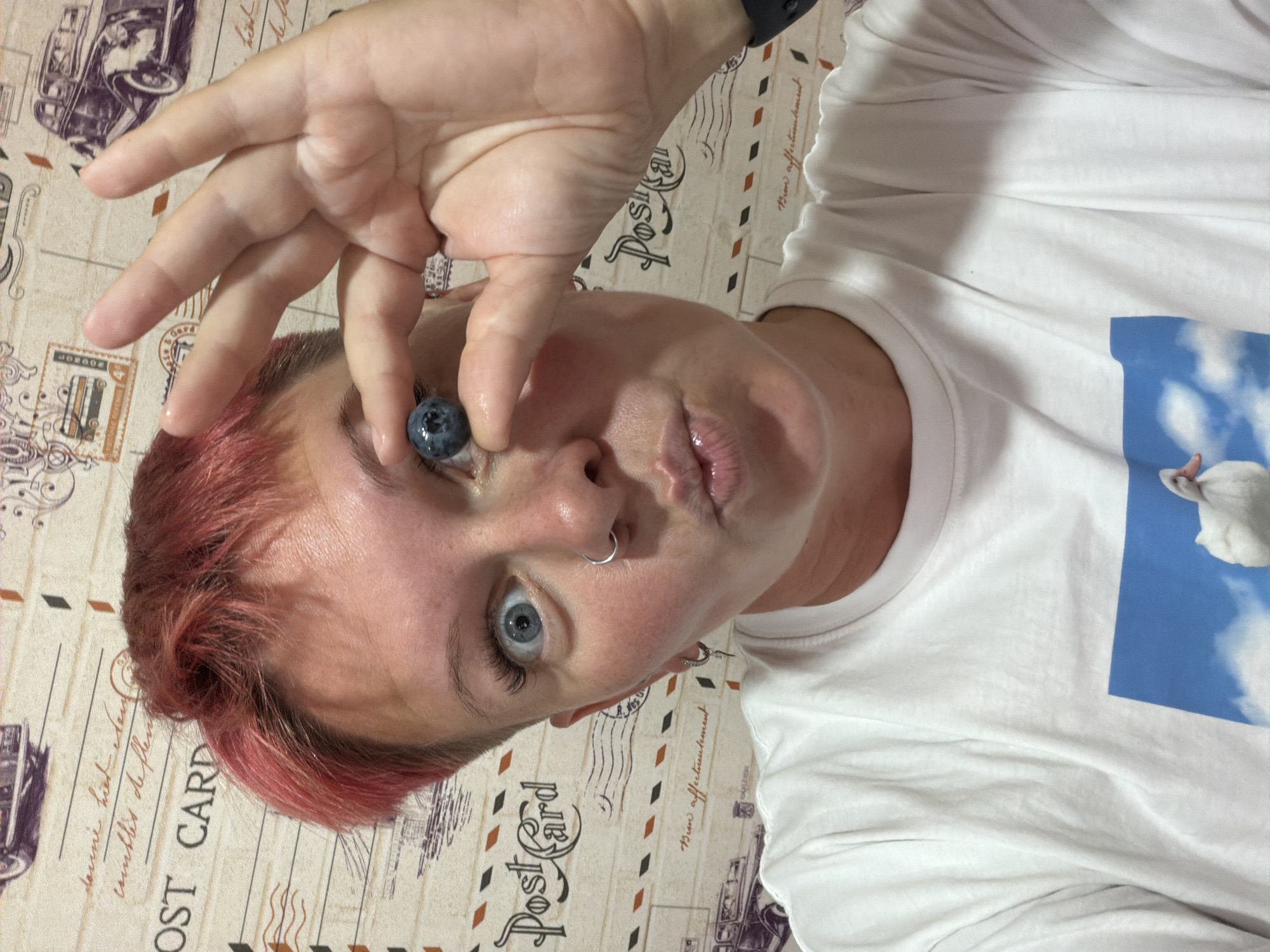 A young woman with short, pink-dyed hair, wearing a white t-shirt, is holding a blueberry to her left eye, covering it. She is looking directly at the camera with a surprised expression and puckered lips. The background is a wall decorated with a postcard-themed wallpaper featuring vintage cars and postage stamps. The lighting is bright, casting soft shadows. The image appears to be a selfie. The location is specified as the city of Chaikovsky, Russia.