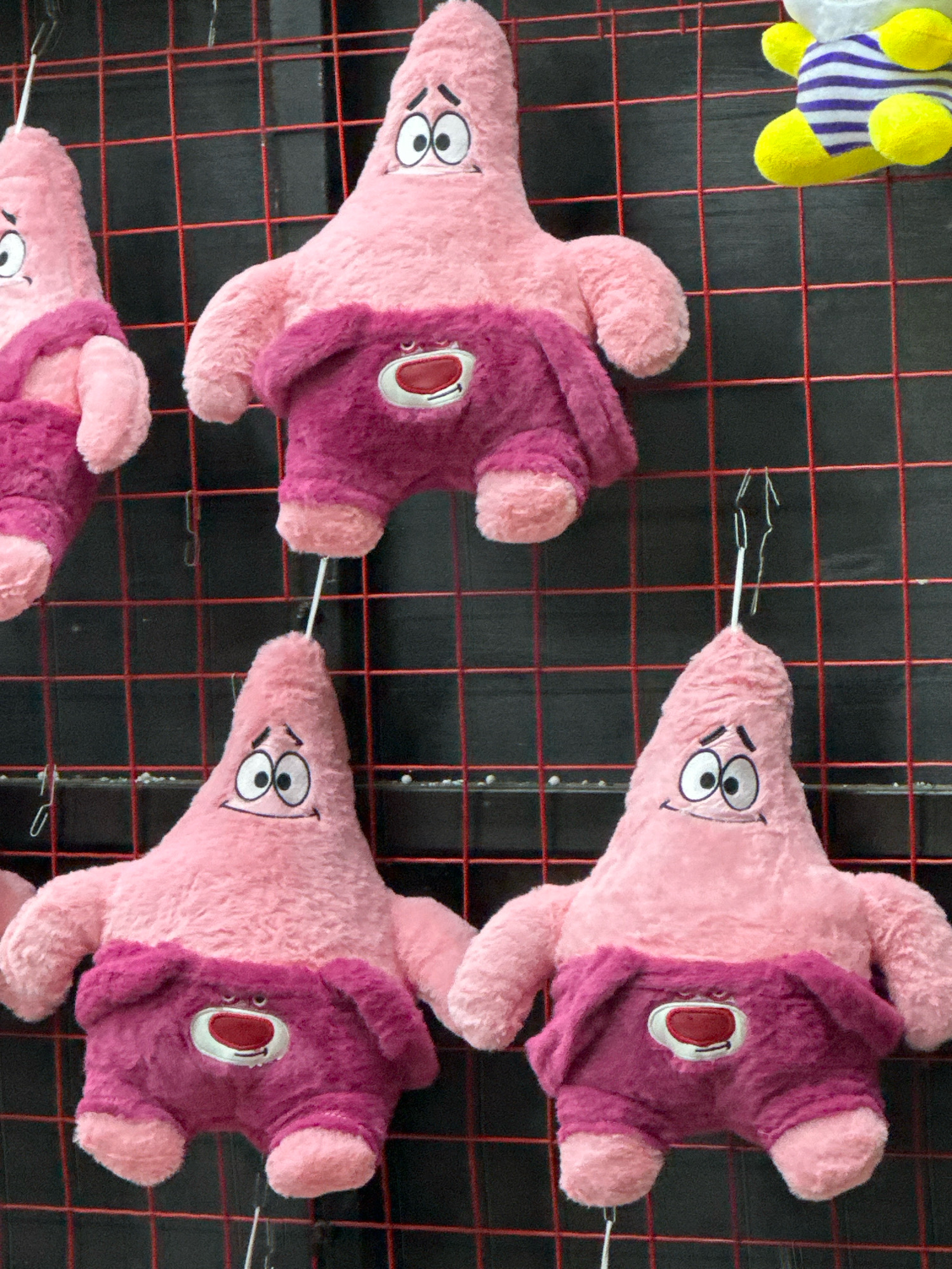 The image shows a close-up of several pink plush starfish toys, resembling the character Patrick Star from SpongeBob SquarePants, hanging on a red grid. Three of the toys are fully visible, and a fourth is partially visible in the upper left corner. The toys are wearing purple shorts with a white oval emblem containing a red circle and black outline. The toys are suspended by thin white strings attached to wire hooks. The background is a black mesh or grid. The scene appears to be a retail display, likely in a souvenir shop or amusement park. There are no people visible in the image. The setting is identified as being in Lermontovo, Russia.