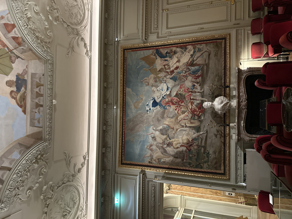This image captures a lavish interior of a grand historic building, specifically identified as likely the Hôtel de Crillon located on Place de la Concorde in the 8th arrondissement of Paris, France.
The room features a high ceiling adorned with intricate white rococo stucco moldings and a central painted fresco depicting classical figures on a trompe l'oeil balustrade, observed from below. A small, white spherical security camera is mounted on the ceiling.
The primary visual focus is a monumental, richly woven tapestry, approximately 4-5 meters wide, framed in gold, which covers a significant portion of the main wall. This tapestry illustrates a chaotic historical or mythological battle scene, featuring numerous figures on horseback and foot engaged in combat, wielding weapons and flags, set against a dynamic sky. Prominent colors include reds, blues, whites, and muted earth tones.
Centered beneath the tapestry is an ornate marble fireplace with a reddish-brown and white veined mantlepiece. Perched on the mantle is a white marble bust of a woman, flanked by two dark, stylized decorative objects.
The foreground contains a seating area furnished with multiple armchairs upholstered in deep red velvet. These chairs are arranged around small tables with dark, reflective surfaces, suggesting a lounge or formal waiting area.
To the left, a partially open doorway leads to an adjoining hallway, revealing marble-clad walls and a green illuminated exit sign. The right side of the room features paneled walls with classical pilasters, consistent with the overall opulent architectural style.