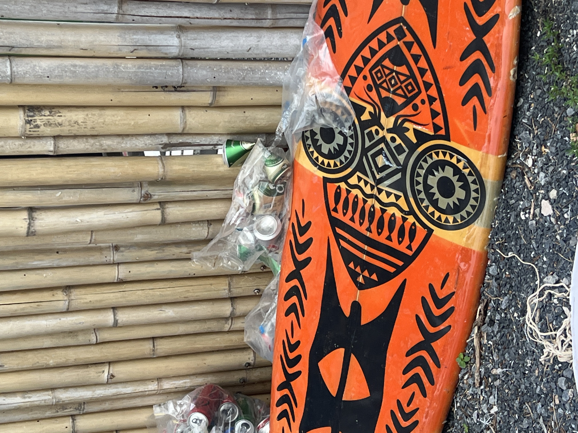 The image displays an outdoor scene featuring a brightly colored surfboard leaning against a bamboo wall. The surfboard, positioned vertically on the right side of the frame, is predominantly orange with prominent black and gold Polynesian-style patterns, including geometric designs, abstract faces, and rows of small fish. It appears to be a functional or decorative piece, showing some signs of use.
Resting between the surfboard and the bamboo wall are several clear plastic bags filled with crushed aluminum cans, indicating collected waste, likely for recycling. Some cans are visibly green, others silver. The bamboo structure consists of multiple horizontal poles, varying in color from light tan to weathered grey. The ground beneath the surfboard and next to the bamboo is covered in dark grey gravel, interspersed with small green weeds or plants. A piece of white string or cord is visible on the gravel in the lower right corner.
No human activity is depicted. The scene suggests a stationary collection point or storage area. The lighting indicates it is daytime, but specific time or weather conditions are not discernible. While some text or branding might be present on the crushed cans, it is obscured and illegible due to the plastic bags and the deformed state of the cans. The setting evokes a casual, possibly tropical or beach-related environment due to the surfboard and bamboo.
