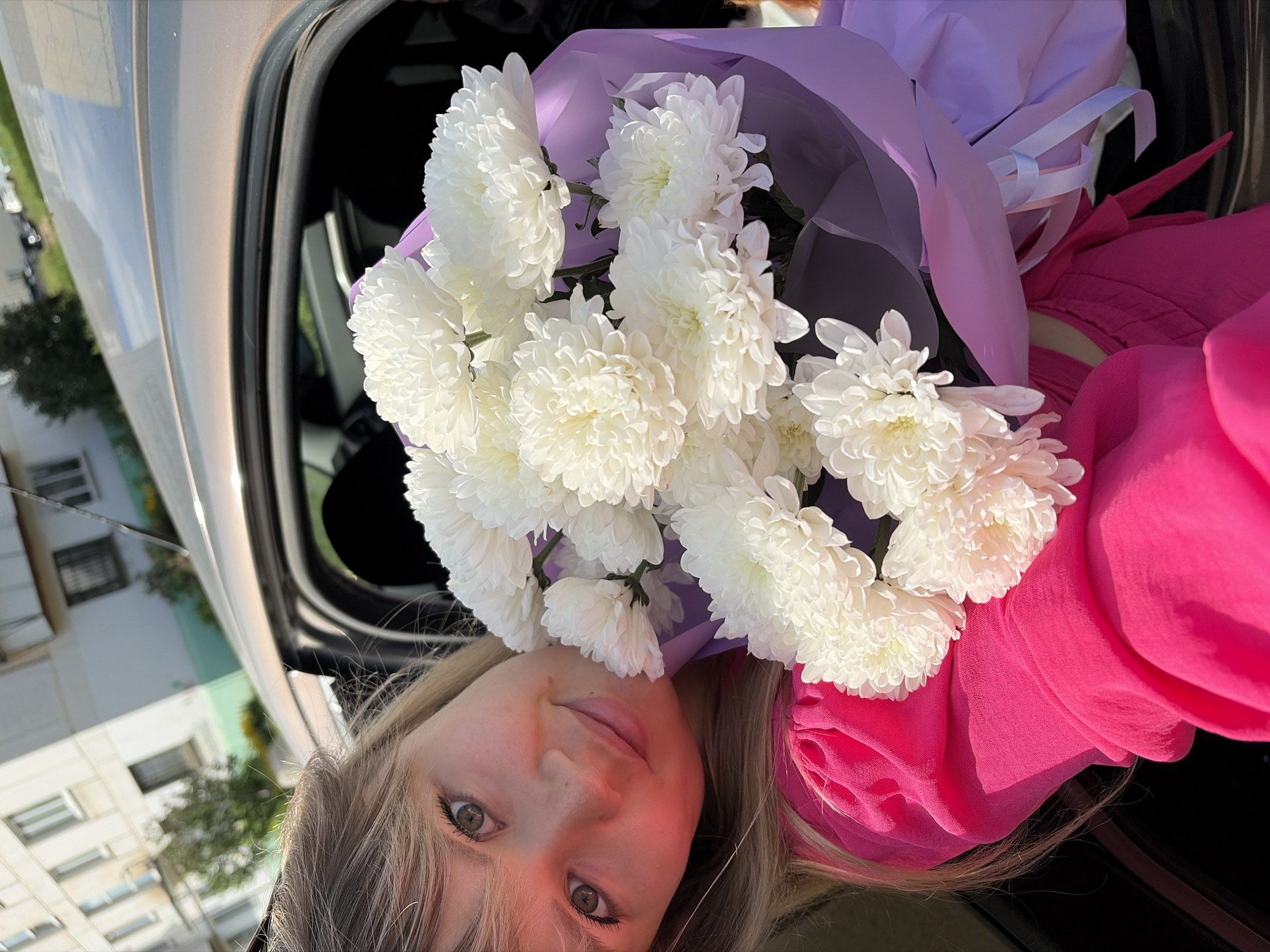 A young woman with blonde hair and green eyes leans out of a car door, holding a bouquet of white chrysanthemums wrapped in purple paper. She wears a bright pink dress with puffed sleeves. The background shows a light-colored apartment building with windows, and some greenery. The car appears to be parked on a street. The scene is set in the city of Chaikovsky, Russia, under a clear sky.