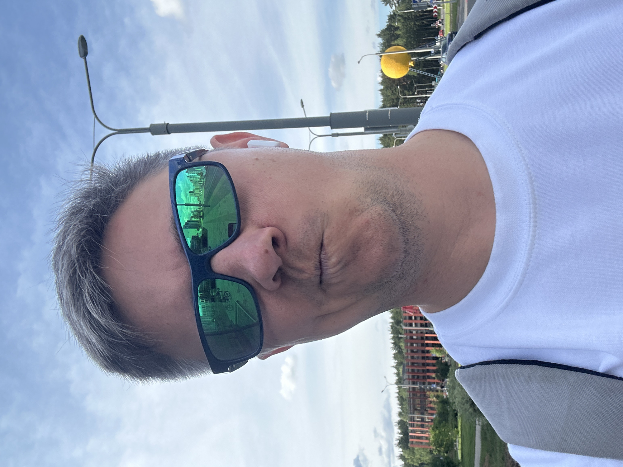 An adult male with short, grey hair and light stubble poses for a selfie outdoors on a partially cloudy day in Бараўлянскі сельскі Савет, Belarus. He is wearing a white crew-neck shirt and a grey backpack, visible on his left shoulder. His lips are pursed in a pouting expression, and his eyes are obscured by dark-framed sunglasses with reflective green lenses. A white wireless earbud is visible in his right ear. The sunglasses reflect a street scene featuring buildings, a road with white markings, and a bicycle. In the background to the left, a multi-story building with a reddish-brown slatted facade is visible behind green trees. To the right, tall grey streetlights with double arms stand against the blue sky, alongside more green foliage. A prominent, large yellow spherical object, possibly a decorative balloon or art installation, is visible on a tall pole in the distance, with what appears to be a ladder leaning against its support structure.
