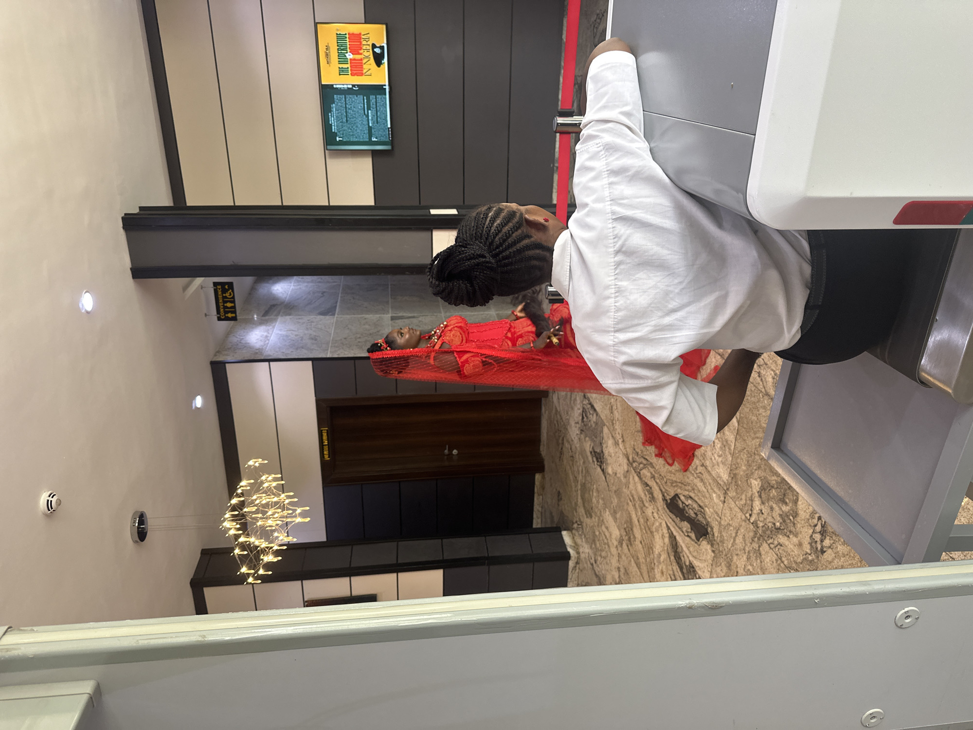 In a modern interior setting, possibly a public building or event venue in Abuja, Nigeria, a young woman adorned in a striking red traditional outfit, including a veil, stands facing left, her gaze directed towards something off-camera. She is positioned near a dark wooden door. In the foreground, a security personnel, facing away from the camera, sits behind a metal detector with a grey surface. They are wearing a white short-sleeved shirt and dark trousers. The background features a television screen displaying what appears to be promotional material, a contemporary chandelier hanging from the ceiling, and signage indicating "CONVENIENCE" with icons for men, women, and accessibility. The floor is a patterned marble.