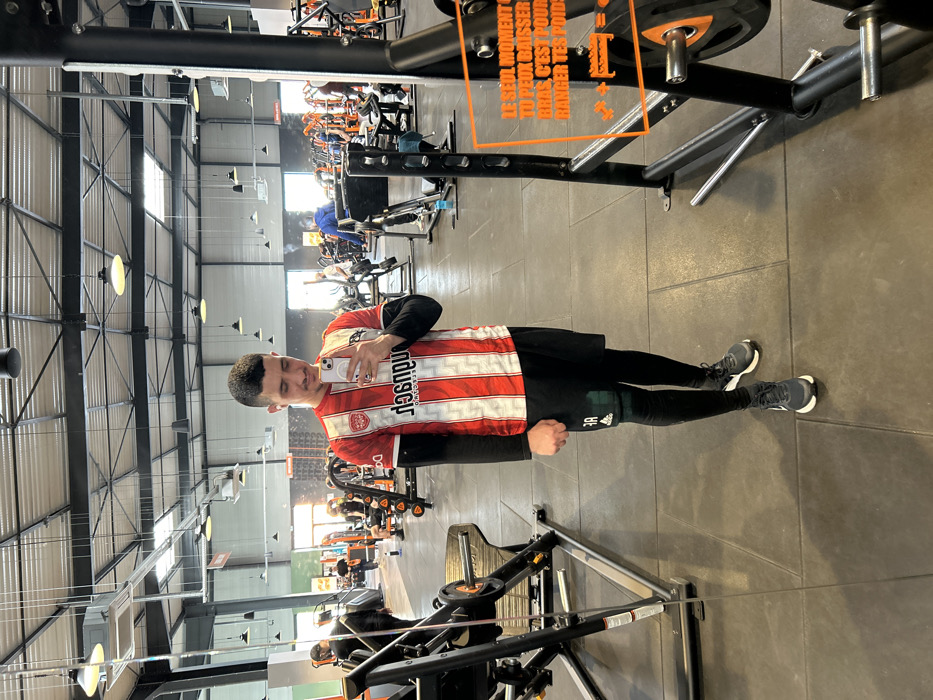 The image displays a full-length mirror selfie of a young adult male inside a Basic-Fit fitness facility, located in Hœnheim, France. The male subject stands centrally, presenting short dark hair, a light mustache, and a goatee. He is dressed in a red and white vertically striped short-sleeved athletic jersey, worn over a black long-sleeved undershirt. Black athletic shorts are worn over black compression leggings, with a green knee sleeve visible on his right leg. His footwear consists of dark athletic shoes with white soles. He holds a white smartphone horizontally with both hands, angled towards the mirror, and exhibits a slight smile. A ring is present on his left ring finger.
The gym floor is constructed of dark gray rubber mats. The facility features a high, open industrial-style ceiling with exposed steel trusses, ductwork, skylights, and numerous suspended light fixtures. Various black and orange Basic-Fit branded fitness equipment is distributed throughout the spacious area. In the background, multiple other individuals are discernible, engaged in diverse exercise routines. To the right of the main subject, a black weight rack system displays an orange translucent sign with French text: "LE SEUL MOMENT OÙ TU PEUX BAISSER LES BRAS, C'EST POUR RANGER TES POIDS" (The only time you can lower your arms is to put away your weights), alongside an illustrative graphic depicting a weight plate and a storage rack.