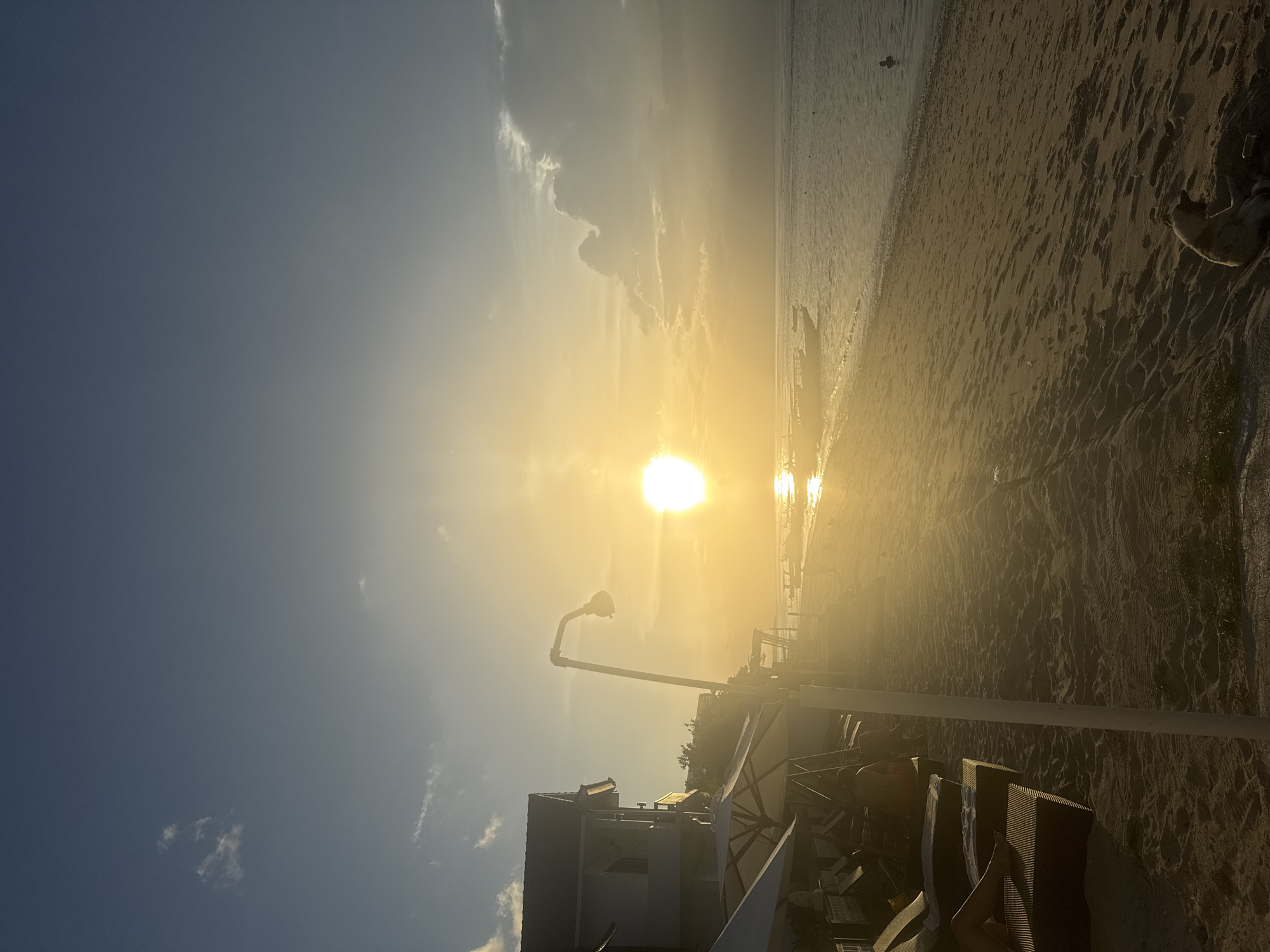 The image captures a beach scene in Nusa Penida, Indonesia, during a vibrant sunset. The sun, a bright orb, dominates the horizon, reflecting strongly on the sand and water. Along the beach, a row of street lights extend towards the horizon. In the foreground, there are various structures that might be part of beachside buildings or amenities, including what appear to be tables, chairs, and part of a building. The beach itself shows visible footprints in the sand, leading towards the water. One person is observed by the shoreline.