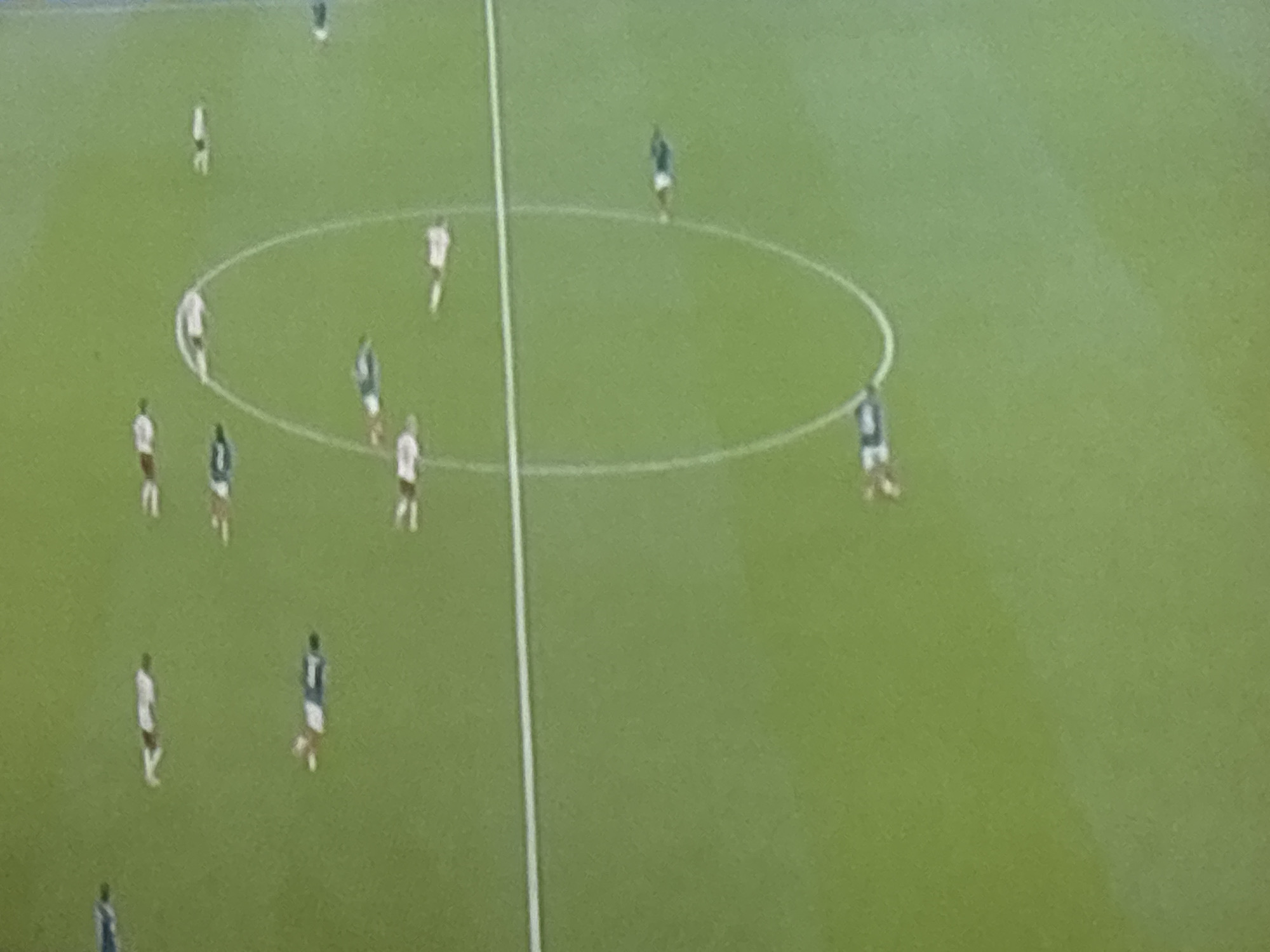 An aerial, wide shot captures a soccer field from a high vantage point, showing the green grass and white markings of the playing area. Several soccer players, dressed in white and blue uniforms, are scattered across the field, some standing and others in motion. A white center circle and a sideline are clearly visible on the grass. The image lacks any identifiable landmarks to pinpoint its location within Sredets, Bulgaria.