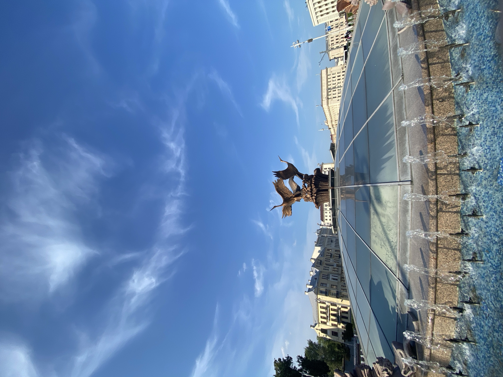 The image captures a bright, sunny day in Minsk, Belarus, likely mid-morning or early afternoon given the clear blue sky with wispy clouds and strong sunlight. The scene is dominated by a modern fountain with a blue tiled basin and a glass enclosure. Water streams from multiple spouts along the edge of the fountain, creating a dynamic visual element.
In the center of the fountain, a bronze sculpture of birds in flight is visible. The birds appear to be swans or cranes, with outstretched wings. Behind the fountain, a row of light-colored, multi-story buildings with visible windows lines a street. The architecture suggests a European city.
The image is oriented vertically, with the sky occupying the majority of the frame. The fountain and buildings are seen from a low angle, emphasizing their scale against the expansive sky. There is no visible text in the image. The overall mood is one of peacefulness and urban beauty on a clear day.
