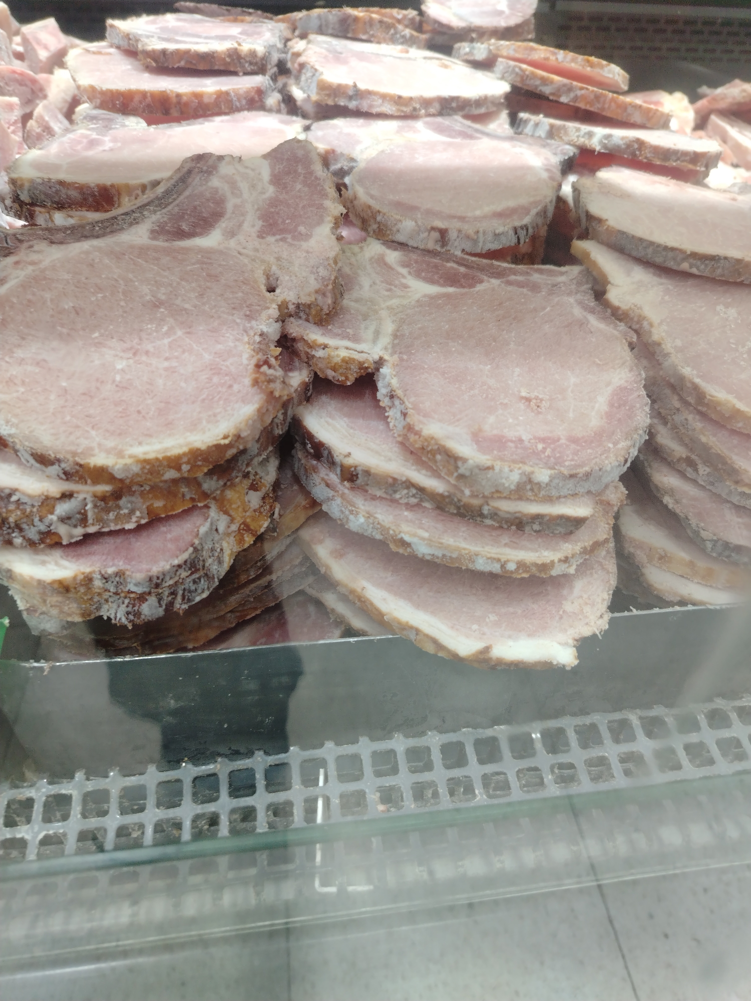 A close-up, eye-level shot shows a display of sliced pork chops in a refrigerated case, likely in a supermarket in Juan Griego, Venezuela. The pork appears to be frozen, with a white frost coating the edges of the cuts, which vary in thickness. The chops are stacked in multiple piles, revealing their pink meat and visible layers of fat. The display case has a metal grate at the bottom, visible through a glass front, and the lighting suggests an indoor commercial setting.