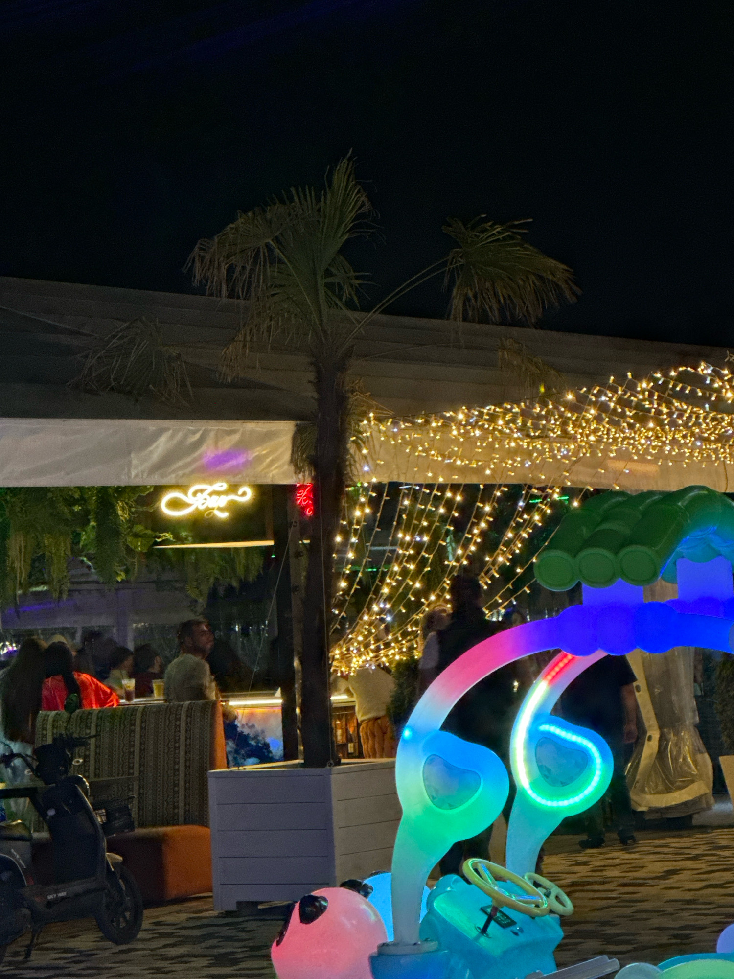 This image depicts an outdoor nighttime scene at a venue in Lermontovo, Russia, likely a bar or restaurant, adorned with numerous string lights. In the foreground, a brightly lit, multi-colored play structure resembling a cartoon car with a steering wheel is visible. To the left, a black scooter is parked. In the midground, several individuals are present, some seated at tables and others standing, appearing to socialize. A neon sign that reads "Bar" in stylized script is illuminated above them. Palm trees are interspersed throughout the scene, contributing to a tropical ambiance. The overall atmosphere suggests a lively and festive evening gathering.