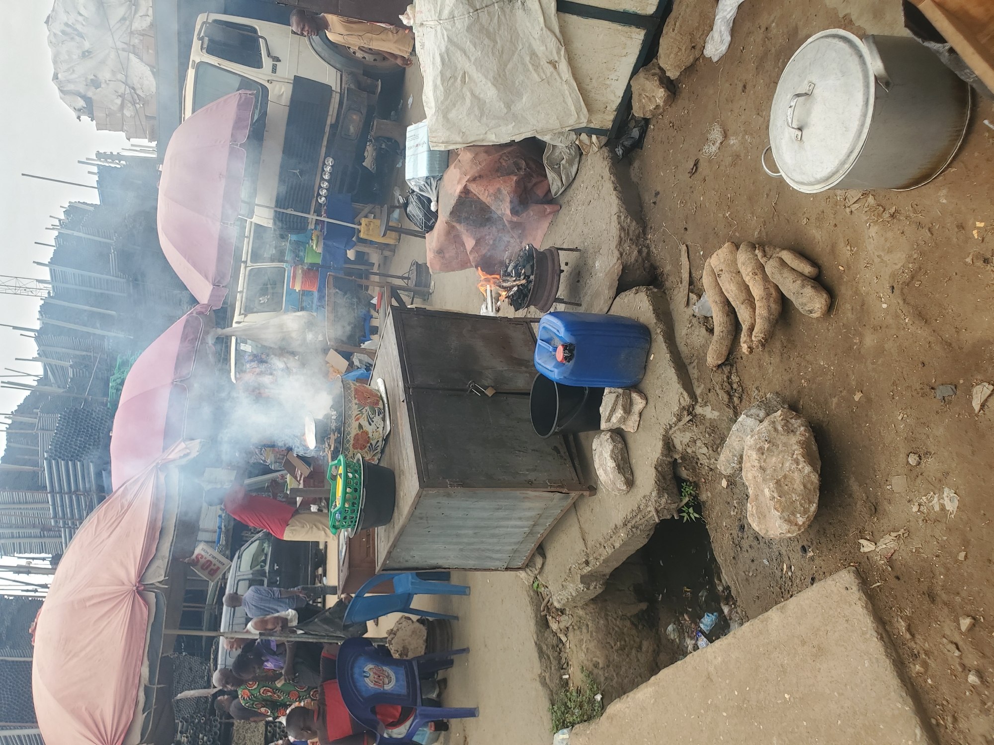 The image depicts an outdoor food stall in what appears to be Fegge, Nigeria. Several individuals are gathered near the stall, seated on blue plastic chairs. A vendor in a red shirt is visible behind a metal table where food is being prepared, with smoke rising from cooking. The stall is shaded by pink umbrellas. A truck is visible in the background. Adjacent to the food preparation area, on the ground, are a large metal pot with a lid, several raw yams, and a stone. A blue plastic container and a black bucket sit on a concrete platform near a small fire in a grill. The ground is dirt and concrete with a drainage ditch. The background shows a metal construction.